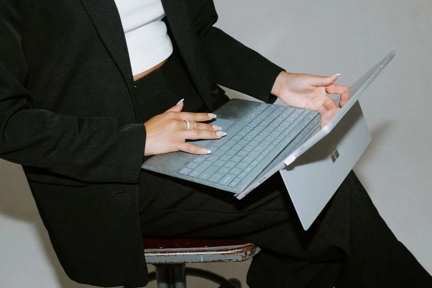 A cropped image of Jaclyn seated on a stool against a plain wall wearing a black suit using a Microsoft Surface device with a keyboard.