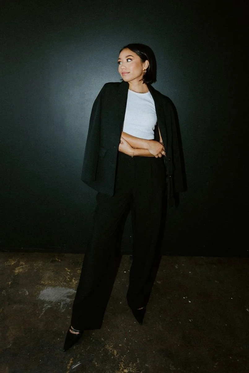 Jaclyn stands arms folded against a black wall, dressed in a white top, black pants, with a black blazer draped over her shoulders, and black high heels. She has short black hair and is looking to her left.