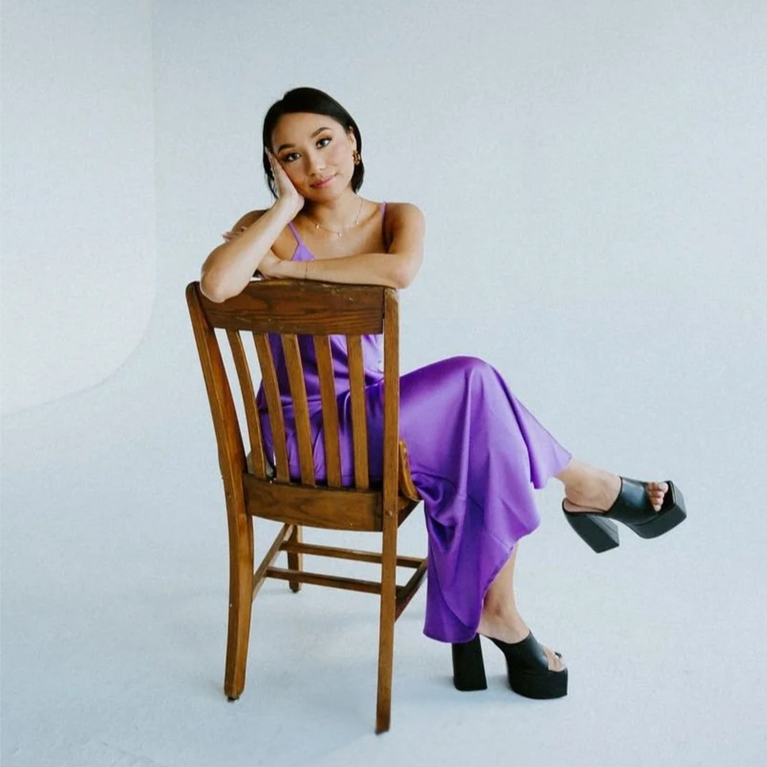 Jaclyn, seated in a wooden chair, wearing a purple dress and her hair short, resting her head on her hand, against a plain white background.