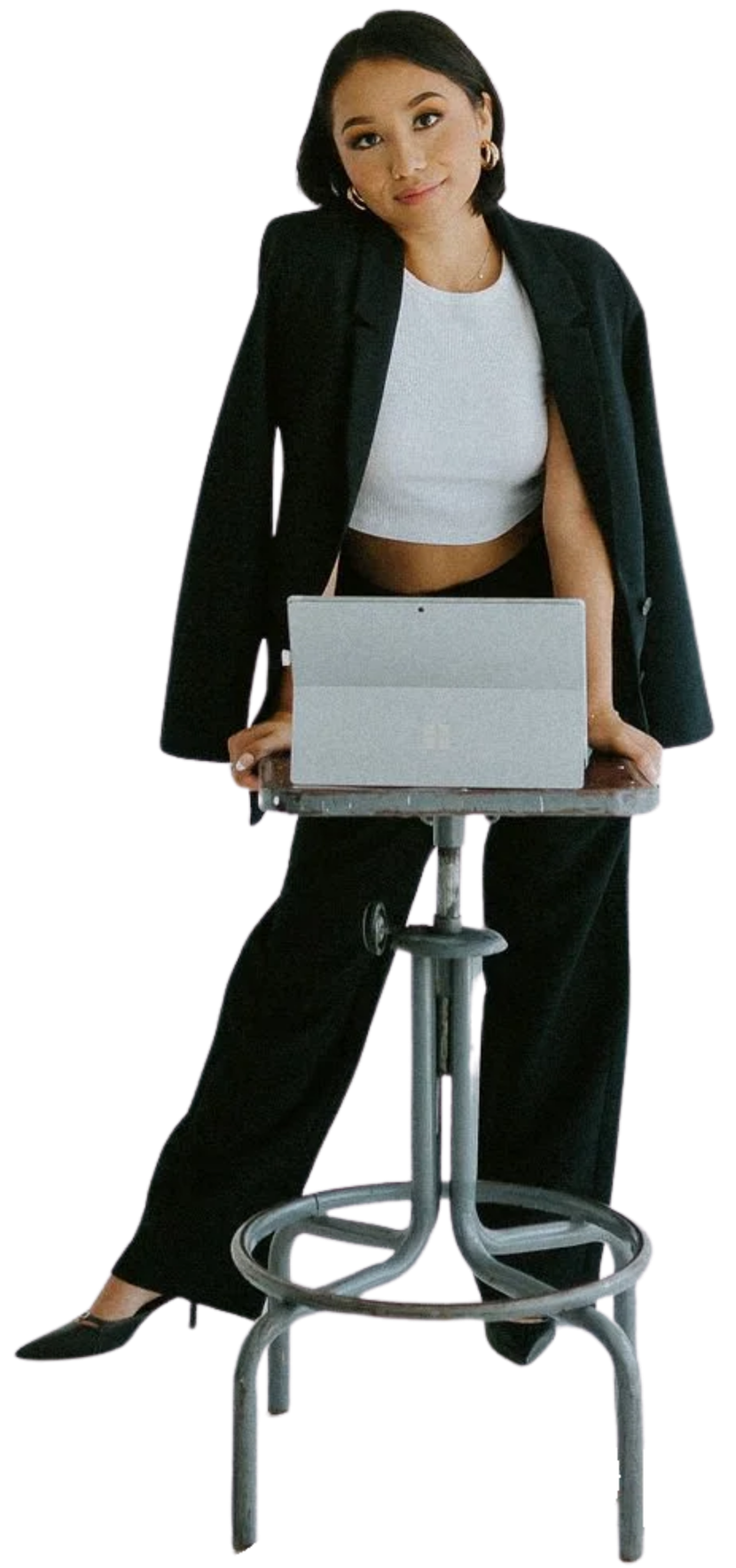 Jaclyn standing behind a stool with a laptop on it, wearing a black blazer, white crop top, and black pants, with a confident expression.