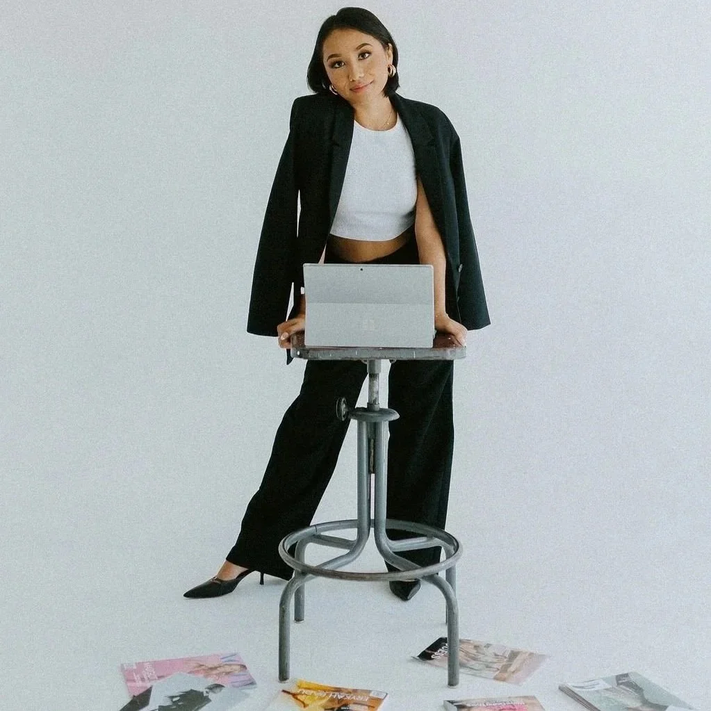 Jaclyn, posed in a black blazer, white crop top, and black slacks standing behind a stool with a laptop on it. She is leaning forward with her hands on the stool, in front of a plain, white backdrop There are magazines scattered on the floor.