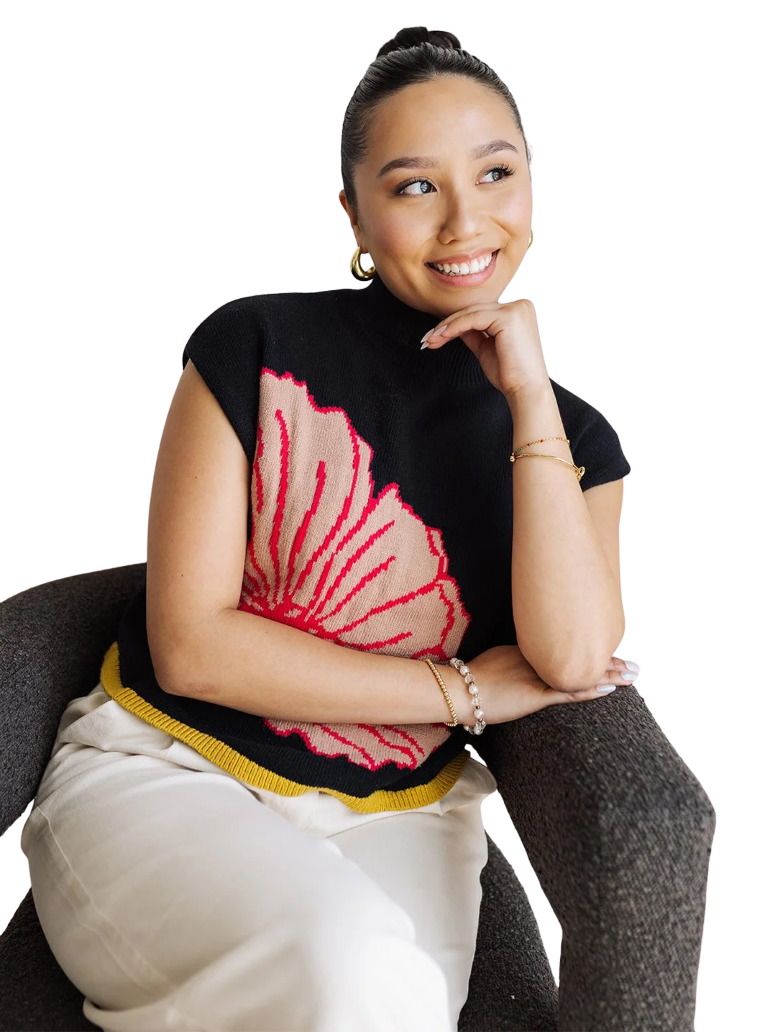 Jaclyn is seated in a chair, arms folded with her chin resting on her hand as she looks to the right. Her hair is slicked back into a high bun. She wears a black top with pink flower and olive trim embroidery, white pants, hoops, a bracelet and ring.