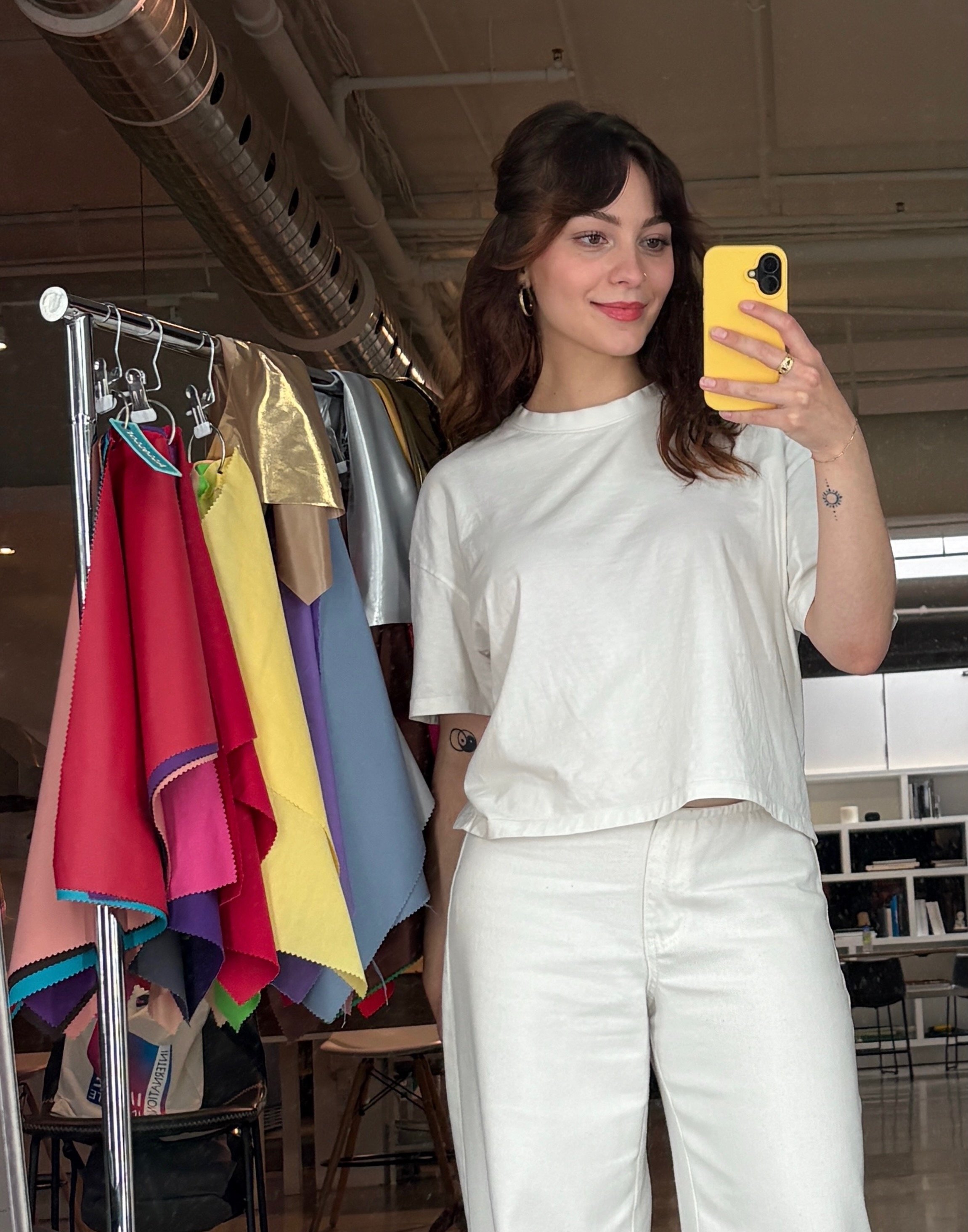 A woman taking a mirror selfie in a clothing store, wearing a white t-shirt and matching white pants, with colorful fabric swatches hanging on a rack beside her.