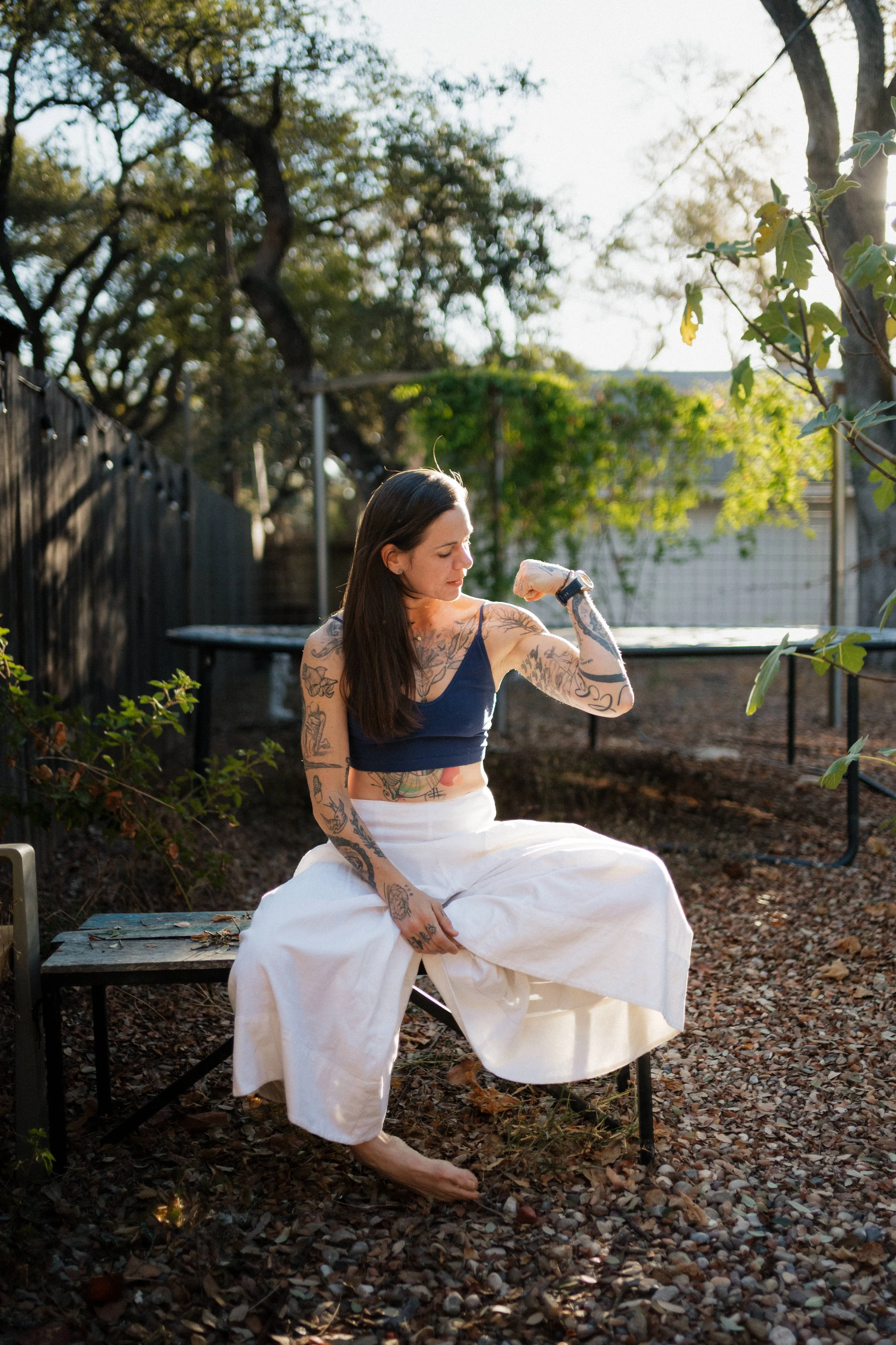 A woman with tattoos on her arms and chest sitting outdoors on a bench, wearing a navy tank top and white wide-leg pants, flexing her right arm to show her bicep, long dark hair, and gazing at her arm with a serious expression, in a backyard with trees, a fence, and a bench, during late afternoon sunlight.