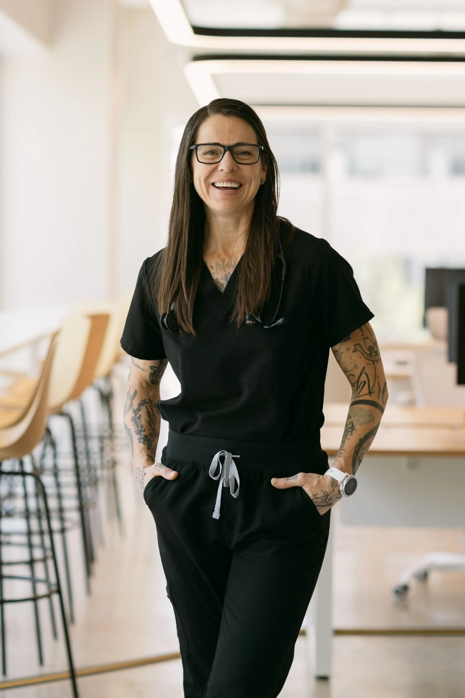 A woman wearing black scrubs, glasses, and a wristwatch, standing in a modern, bright room with a smile, hands in her pockets, and tattoos on her arms.