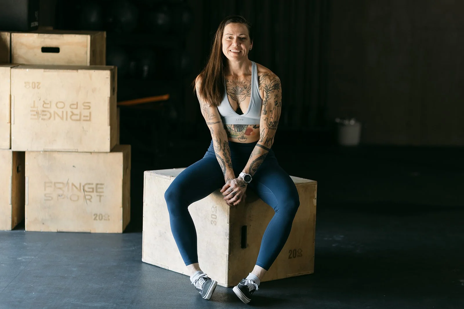 A woman with tattoos sitting on a wooden box in a gym, smiling, wearing a sports bra, leggings, and sneakers.