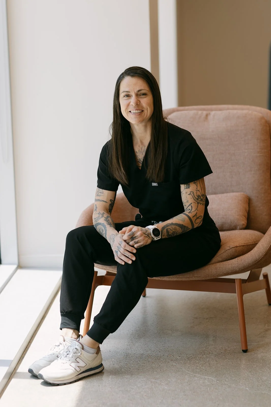 Woman sitting in a tan armchair with tattoos on her arms, wearing black scrubs, white sneakers, and a smartwatch, smiling in a well-lit room.