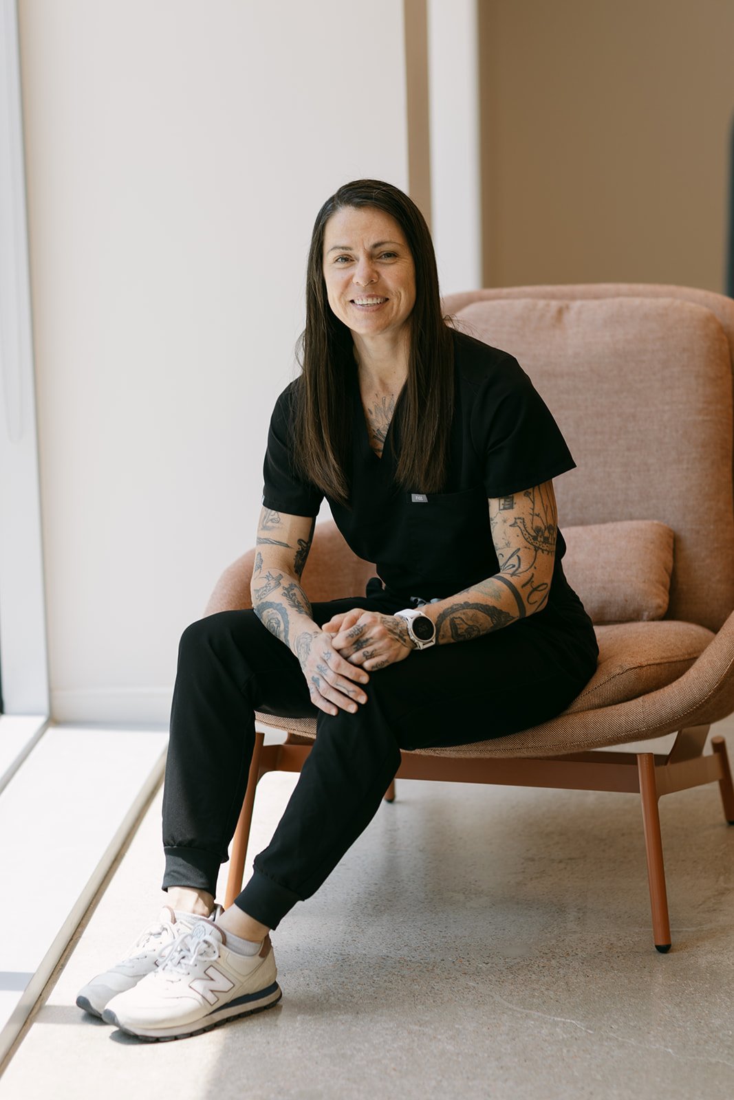 A woman with long dark hair, tattoos on her arms, wearing black scrubs and white sneakers, sitting on a pink armchair in a well-lit room.