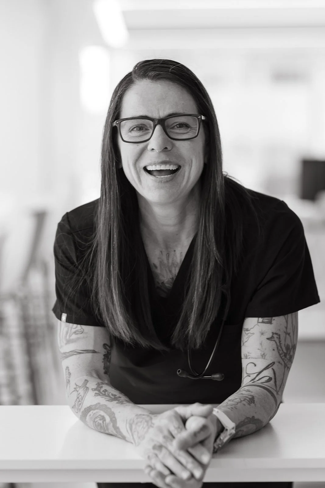 A woman with long dark hair, wearing glasses and a black shirt, smiling while sitting at a table in an indoor setting.