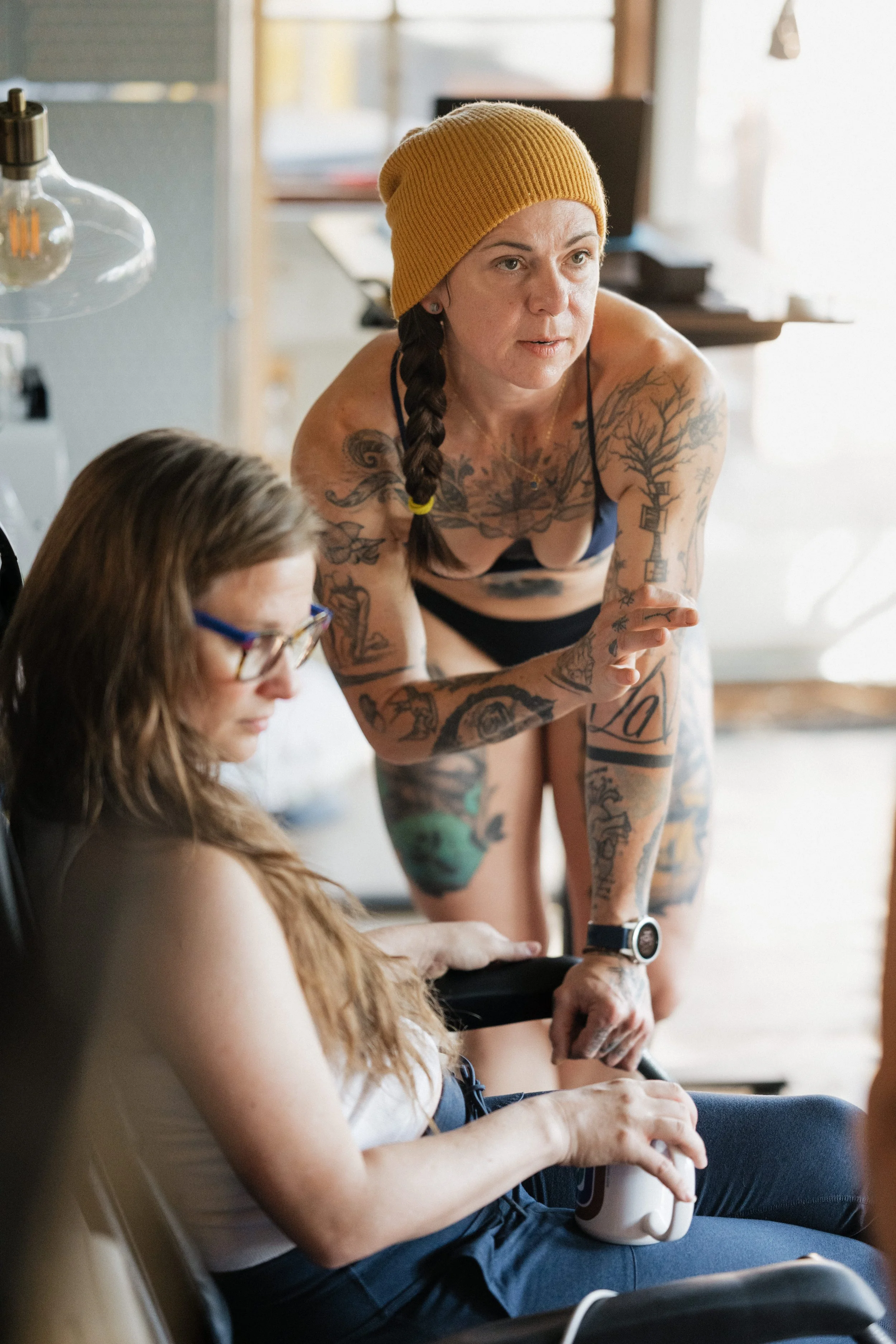 A woman with tattoos, wearing a yellow beanie, leaning over and talking to a seated woman with glasses, who is holding a mug, in a bright room.