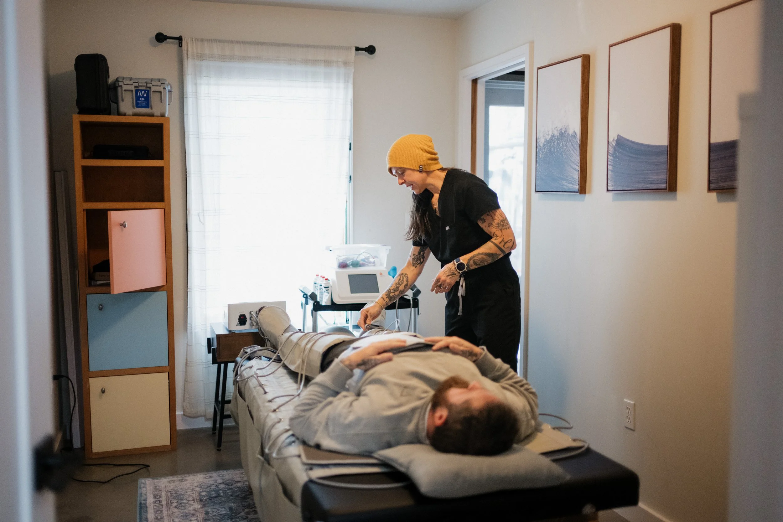 A nurse or caregiver attending to a patient in a hospital room, lying on a bed with medical monitoring equipment nearby. The caregiver is wearing a yellow beanie and has tattoos on her arms.