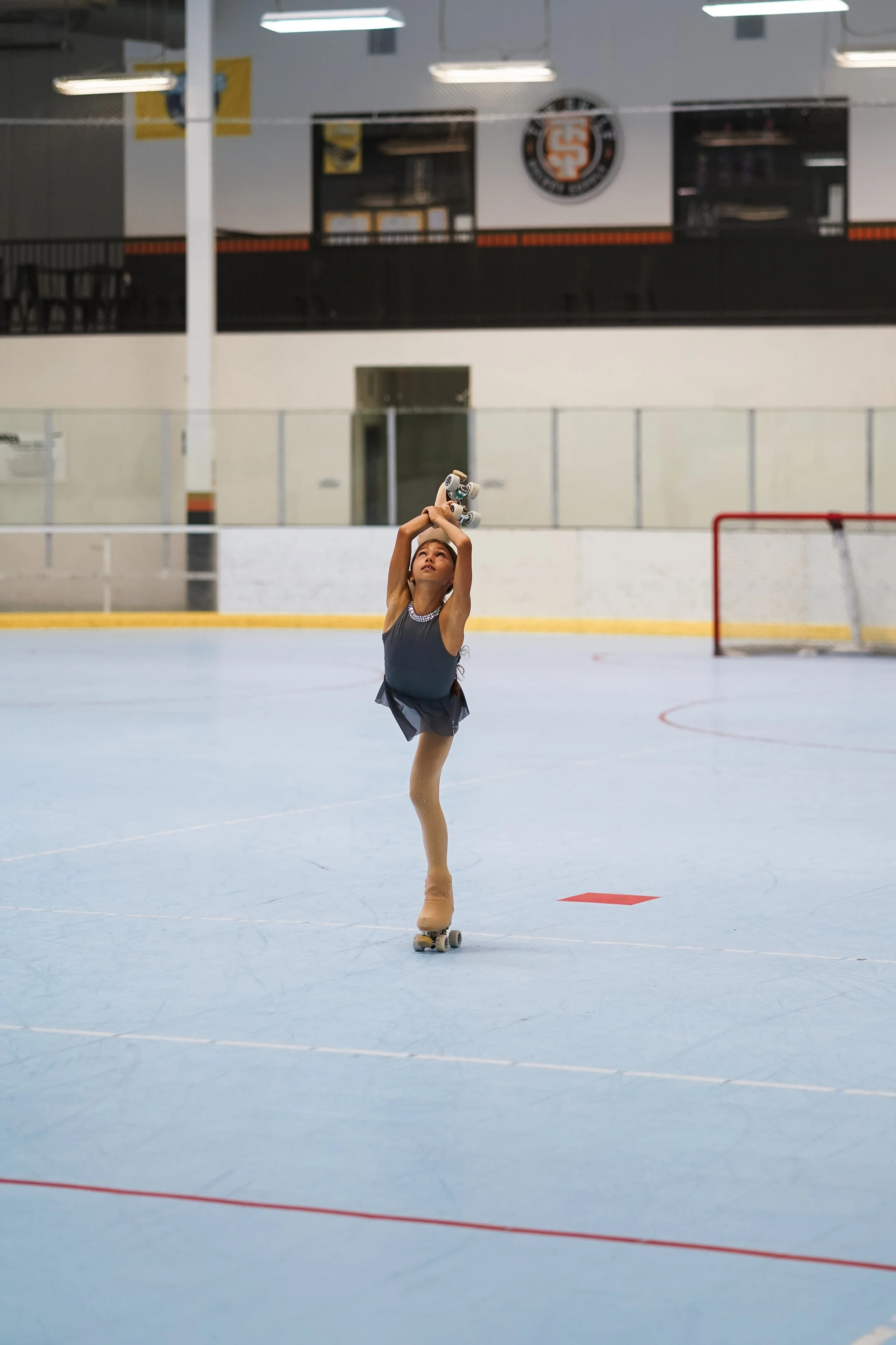 A young girl on roller skates practicing on an indoor ice rink, holding a skate with one hand raised above her head, wearing a grey dress, with a hockey goal and rink boards visible in the background.