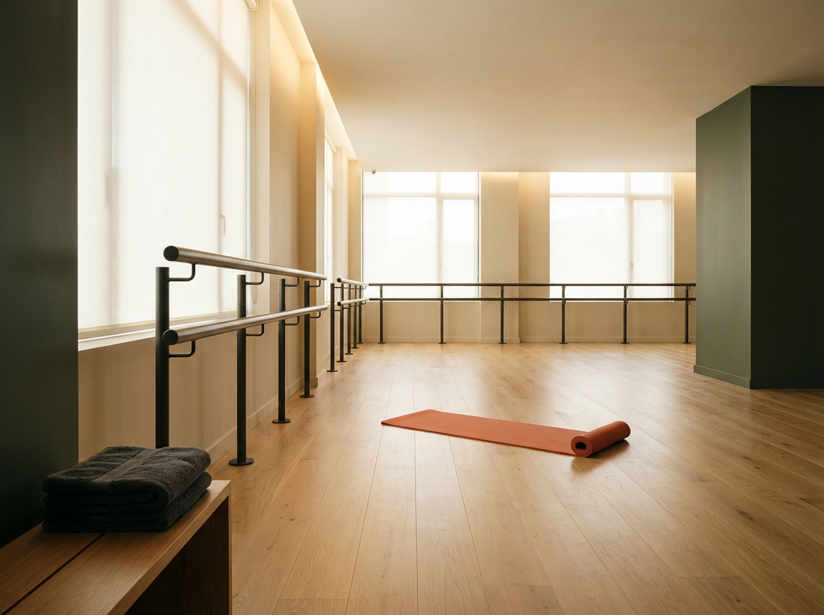 An empty dance studio with large windows, a wooden floor, a rolled-up yoga mat on the floor, and folded towels on a bench.