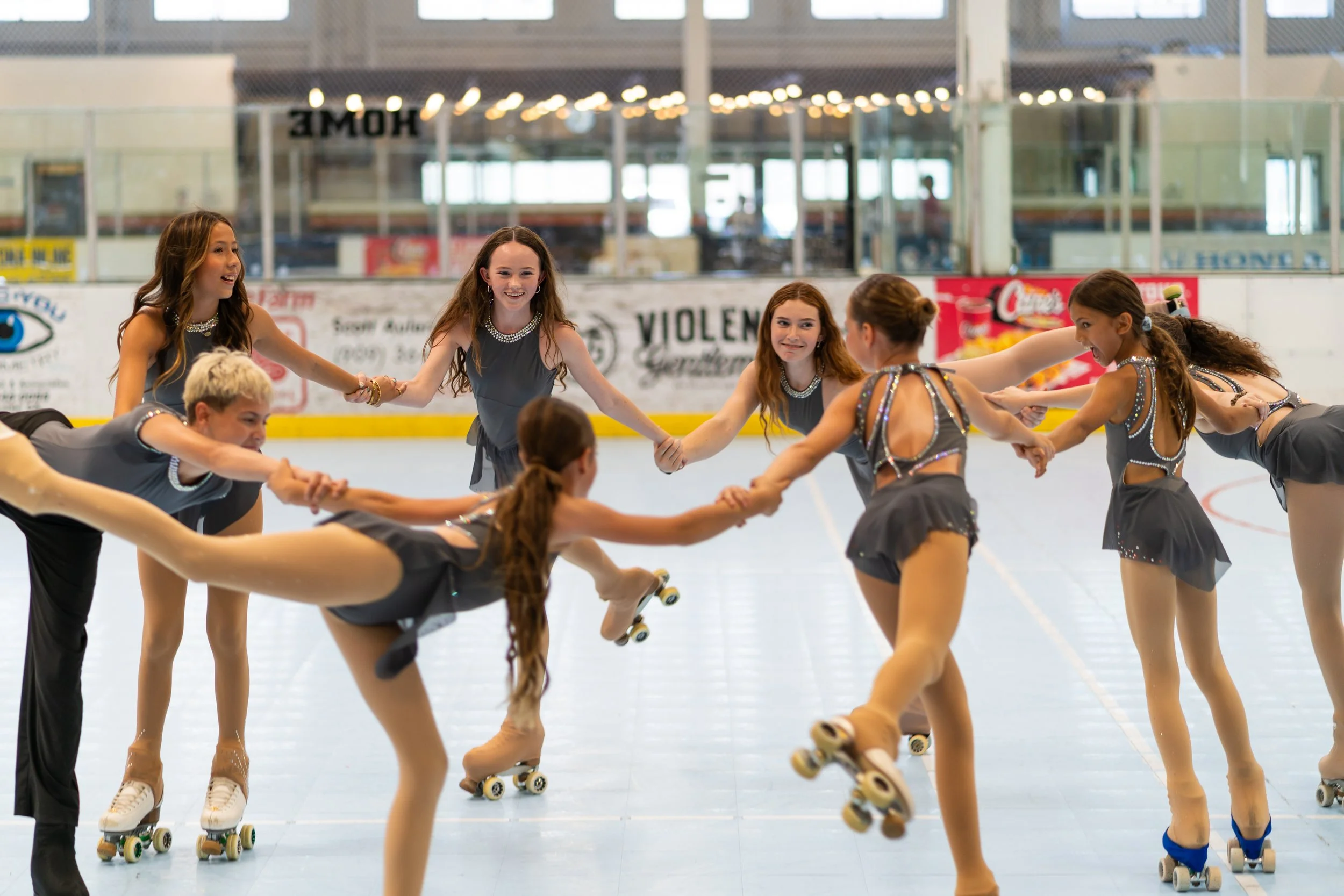 A group of young girls in matching dance costumes holding hands on roller skates in an indoor ice skating rink.