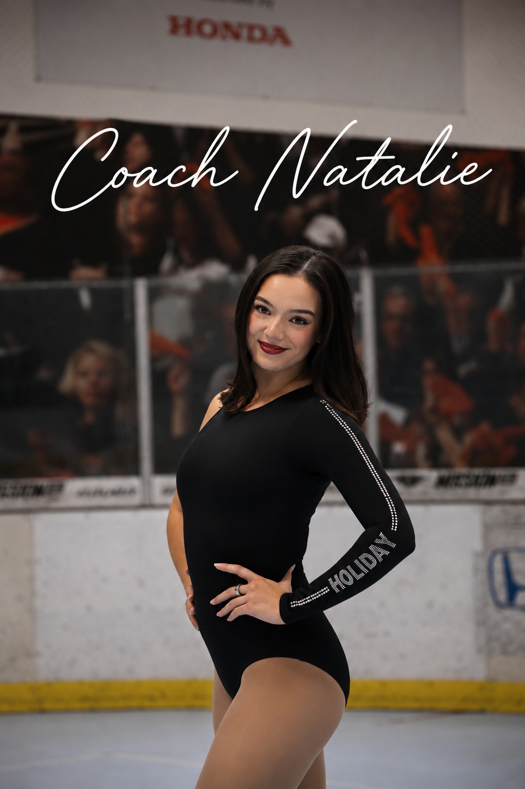 A young woman with black hair, wearing a black dance leotard with 'HOLIDAY' printed on the sleeve, standing in an indoor arena with a hockey rink and a crowd in the background, smiling at the camera. Text overlay reads 'Coach Natalie'.