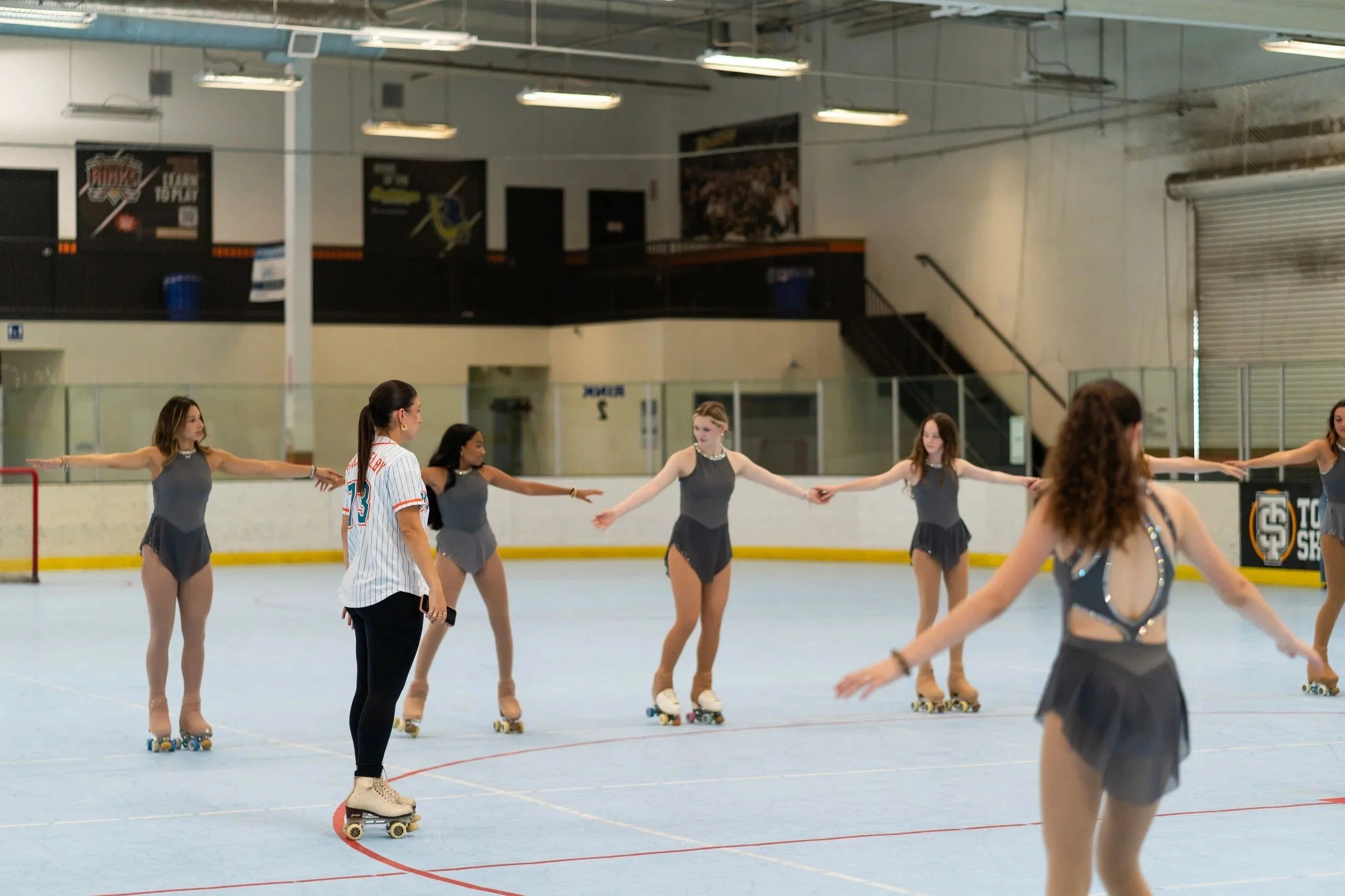 A group of young women in figure skating practice on an indoor ice rink. They are dressed in matching gray costumes, and some are wearing roller skates. An instructor, dressed in a baseball jersey and black pants, is standing among them, giving guidance. The background shows black and white walls with banners and a staircase.
