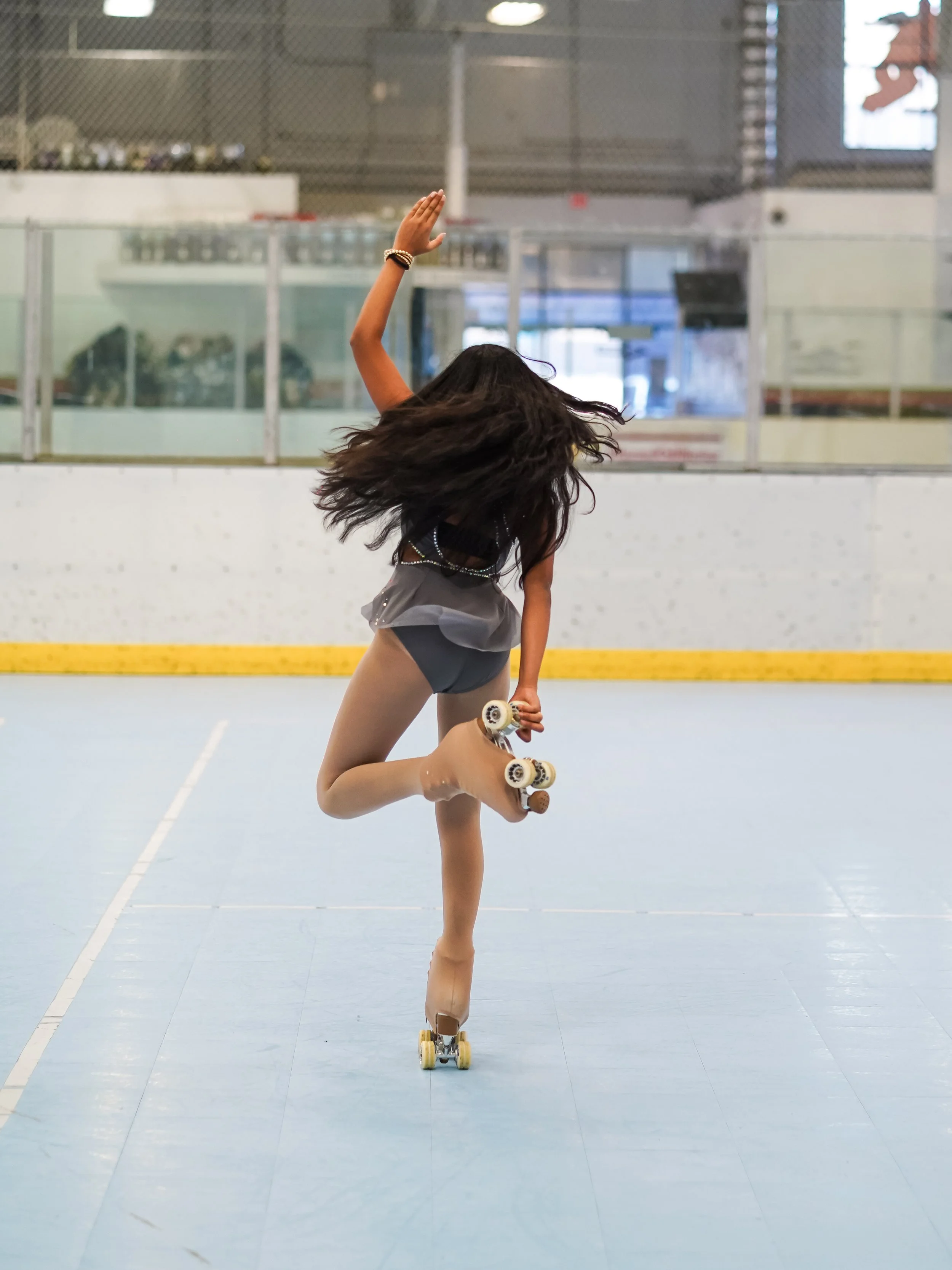 A young girl in a black outfit with a short skirt is roller skating on an indoor rink. She is balancing on one leg with her other leg bent backwards and her arm raised in a skating pose.