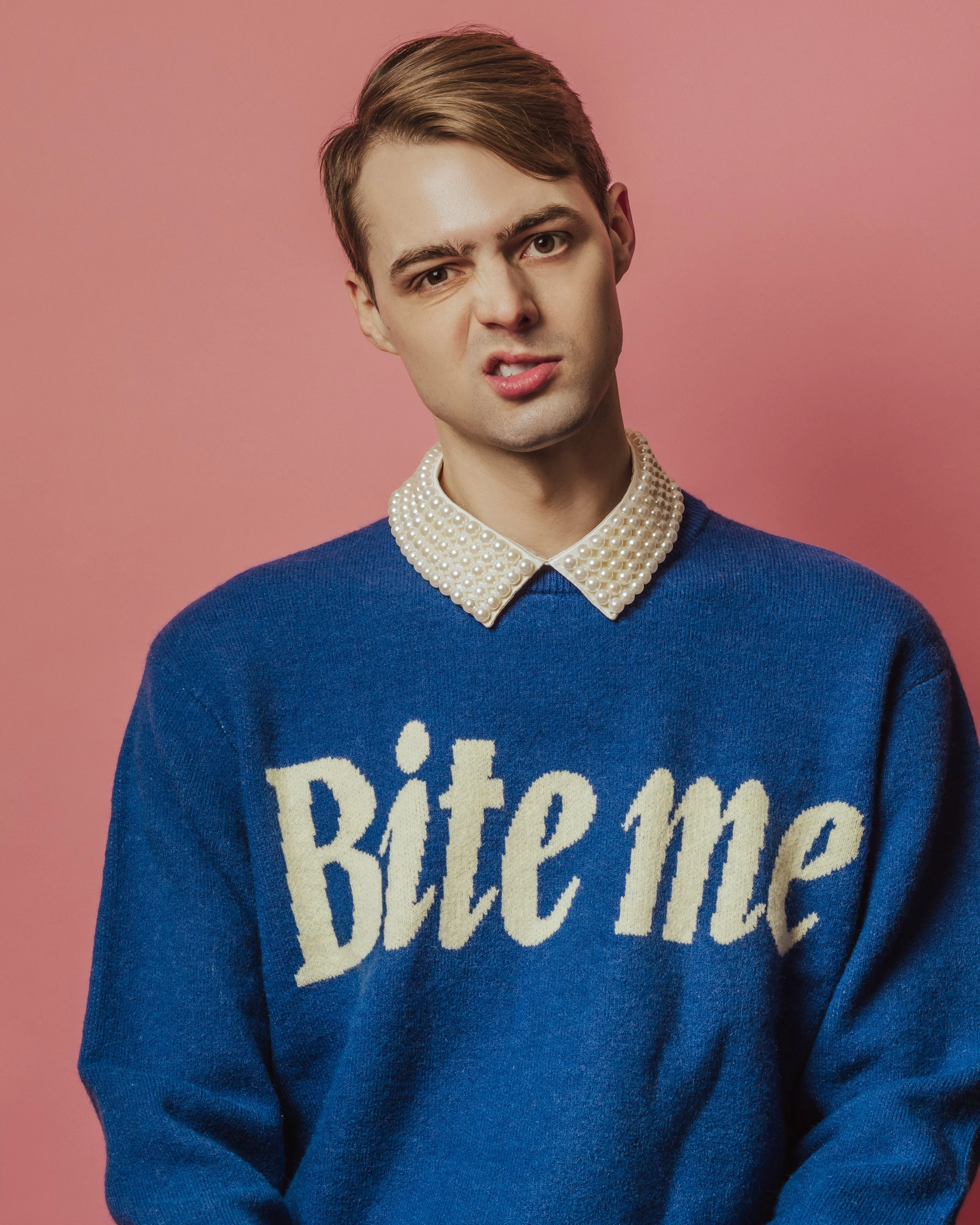 Young man with light skin and short brown hair, wearing a blue sweater with a white pearl collar, standing against a pink background, making a facial expression with furrowed eyebrows and slightly parted lips.