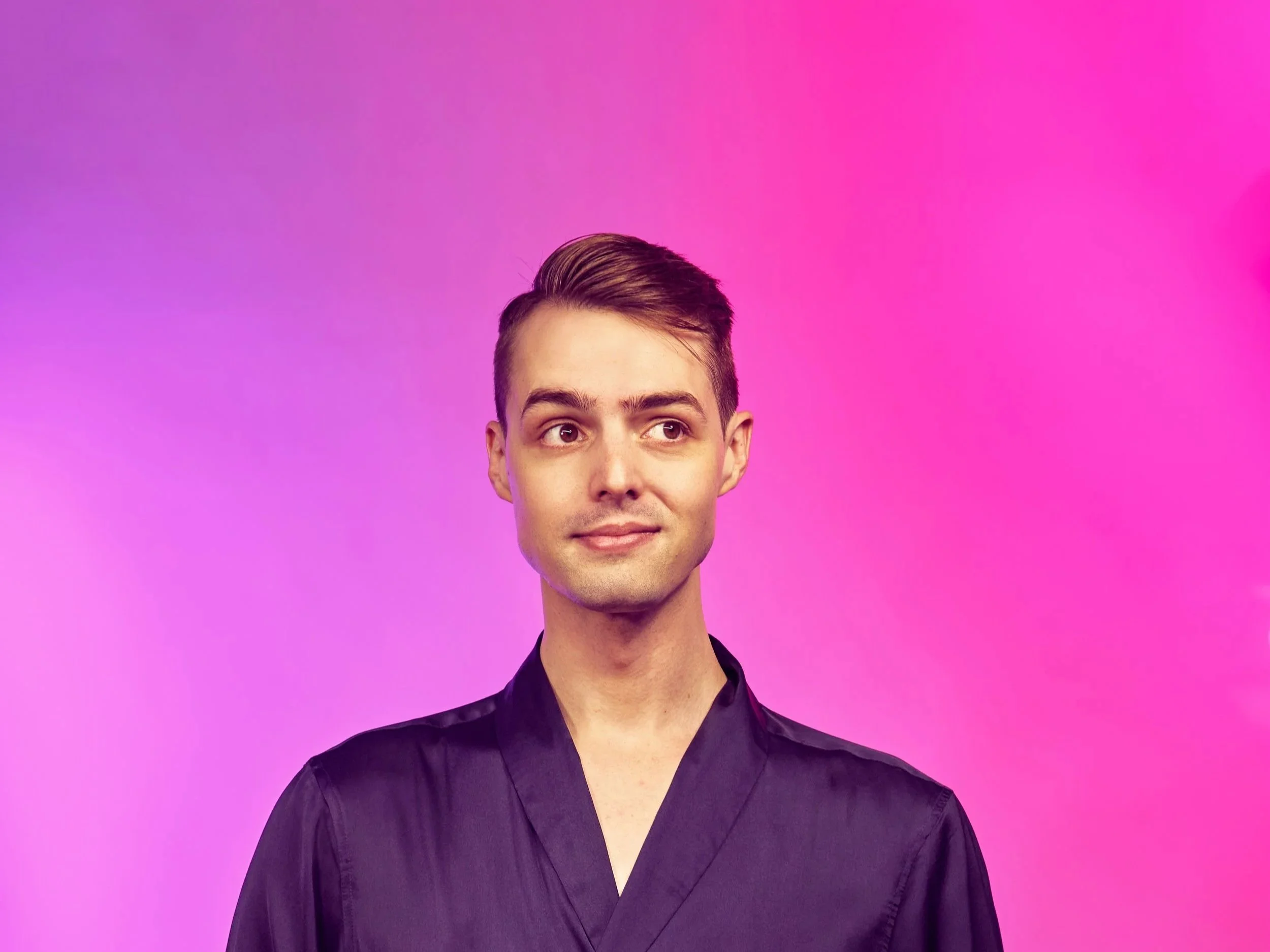 Portrait of a young man with dark hair wearing a black shirt against a pink and purple gradient background.