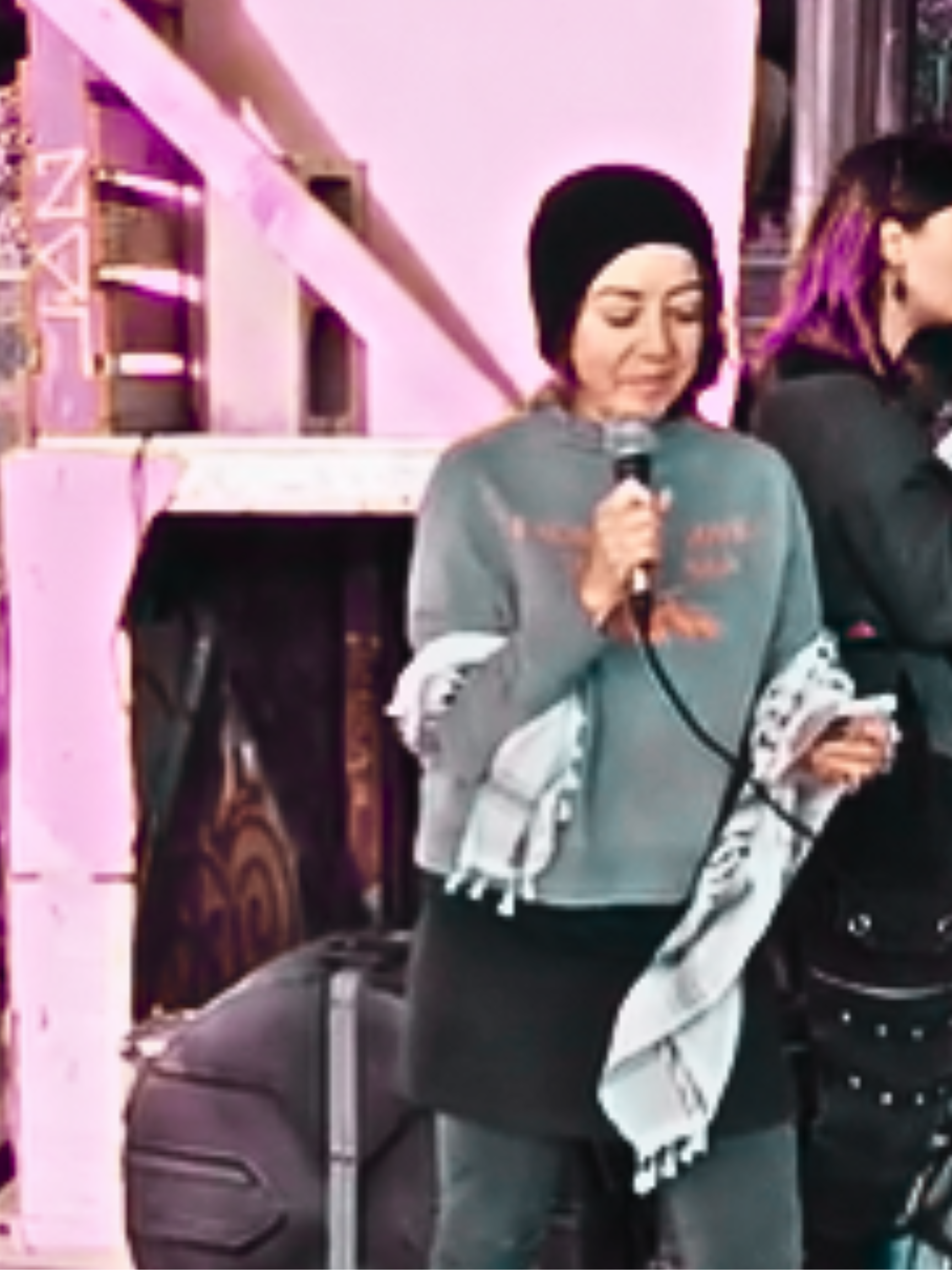 Two women standing outdoors near a pink building, one holding a microphone, and wearing a black beanie hat and a black and white Palestinian keffiyeh.