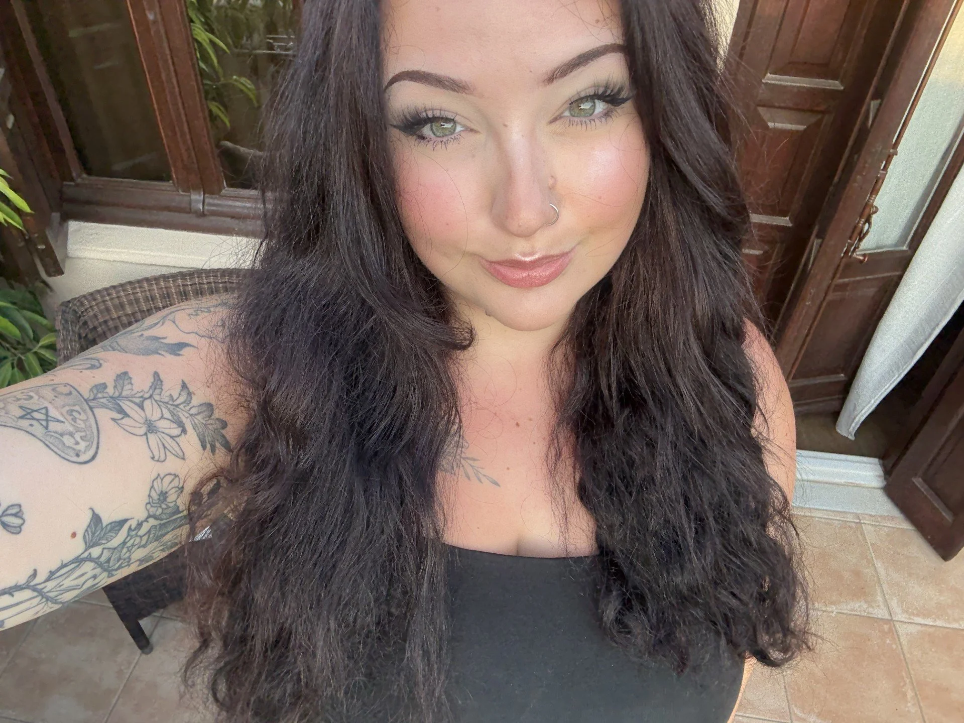 A woman with long dark hair and green eyes, smiling, wearing a black top and sporting tattoos on her left arm, sitting indoors near a wooden door and window.