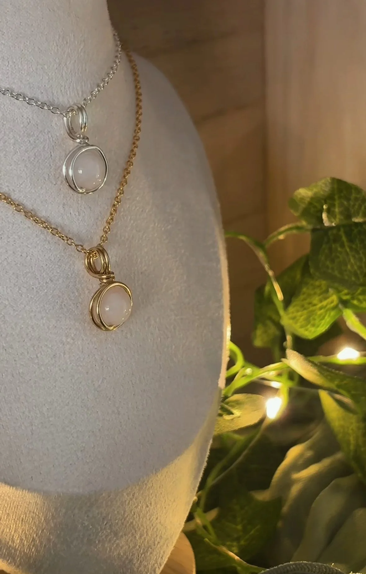 Close-up of a dual-layer silver and gold chain necklace with circular pendants on a white bust display. Green leafy plant with string lights is in the background.