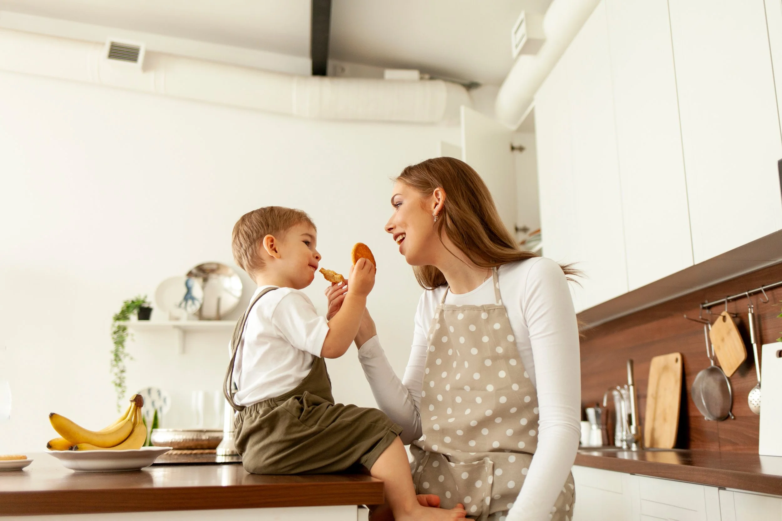 mother in apron feeds child in the kitchen, young woman cook gives cookies to her little son to eat at home By Богдан Маліцький