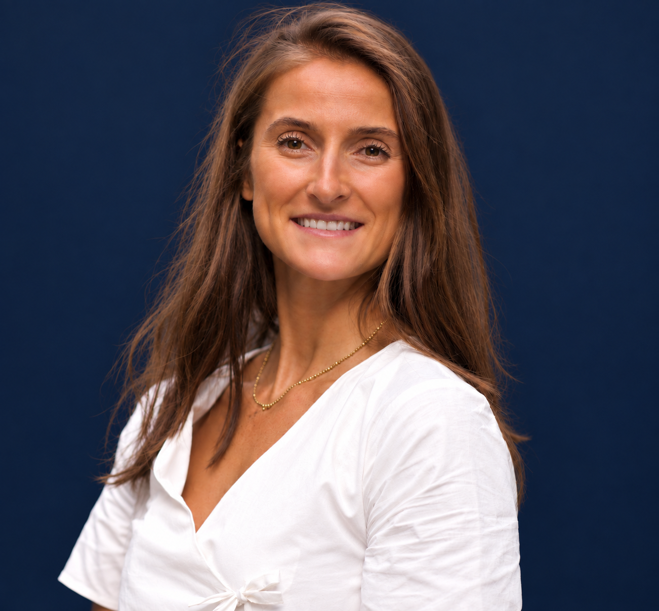 Portrait d'une femme souriante aux cheveux bruns, portant une chemise blanche et un collier doré, avec un arrière-plan bleu foncé.