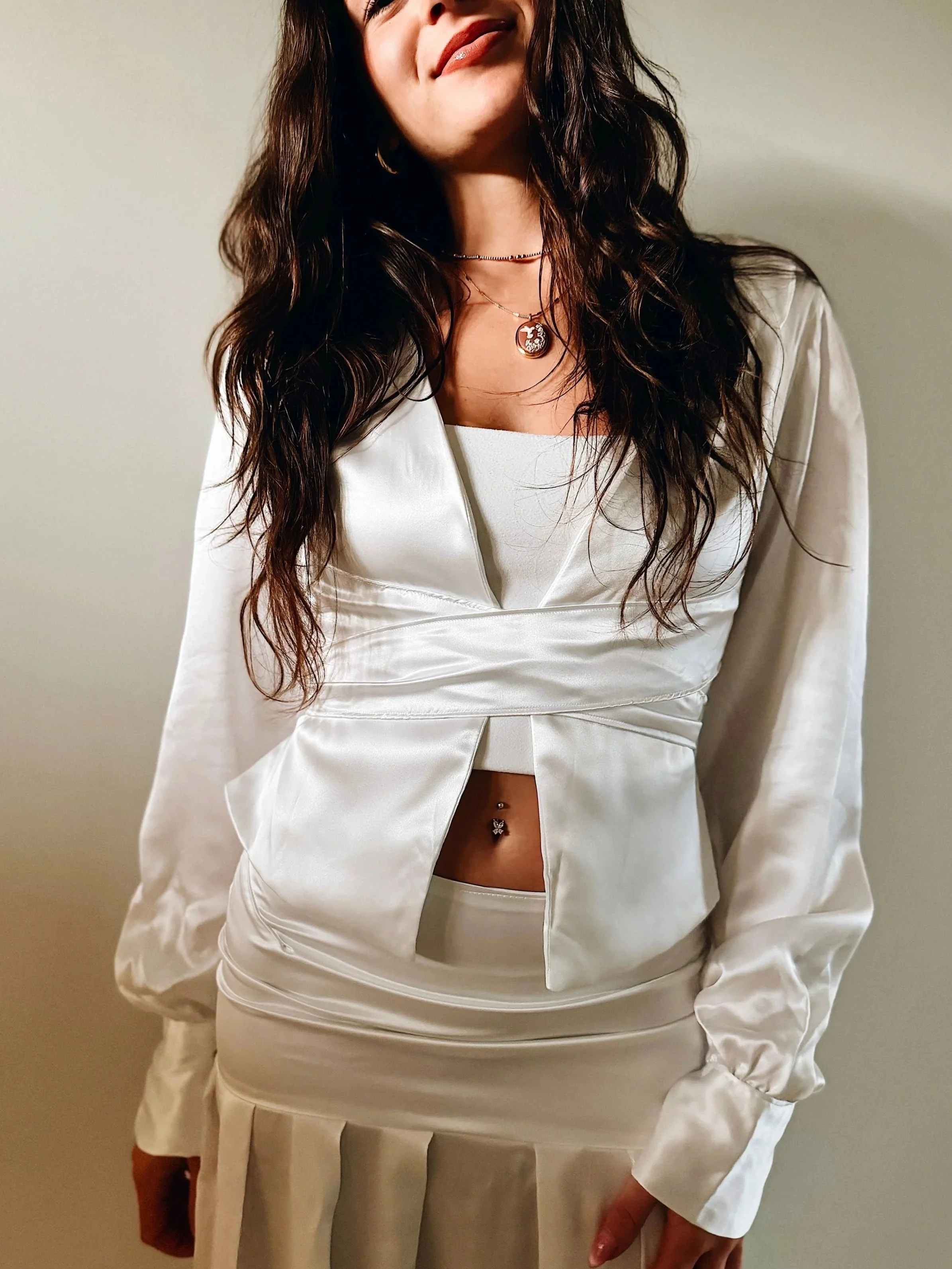 A woman with long wavy dark hair wearing a white satin blazer and matching skirt, adorned with layered necklaces, including a small round pendant and a choker, standing against a plain wall.