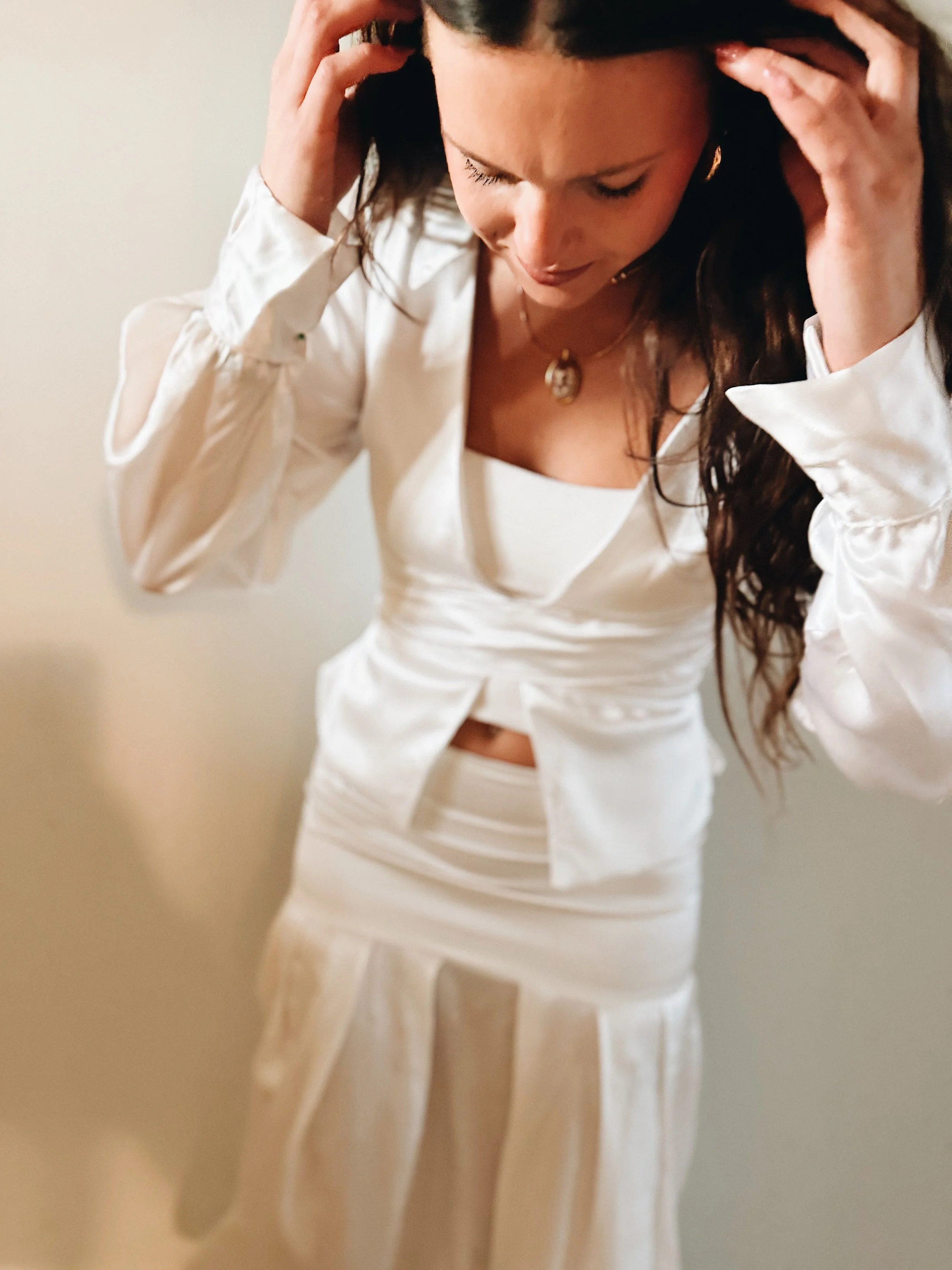 A woman wearing a white satin blazer and matching skirt, with dark hair, holding her head with both hands, looking downward with a distressed expression.