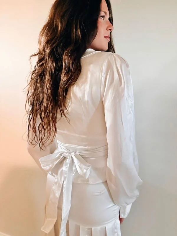 A woman with long wavy brown hair wearing a shiny white satin dress with a waist bow, standing sideways and looking to the right.