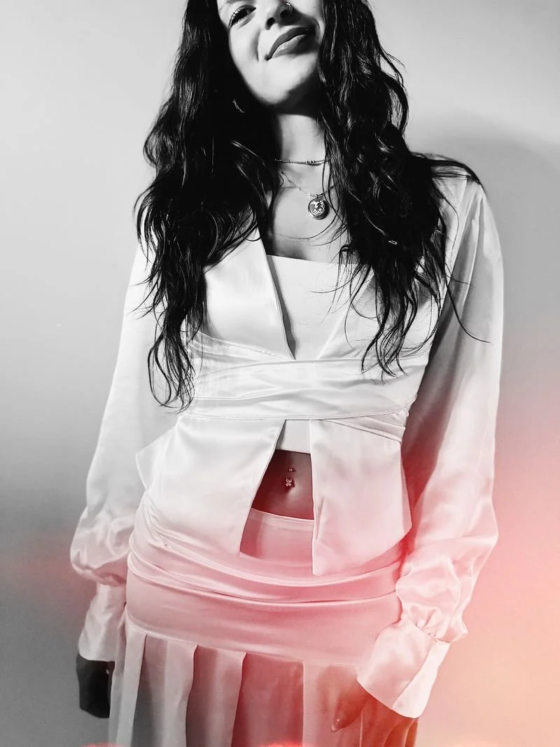 Black and white photo of a smiling young woman with long wavy hair, wearing a satin blazer and skirt, a layered necklace, and visible belly button piercing.