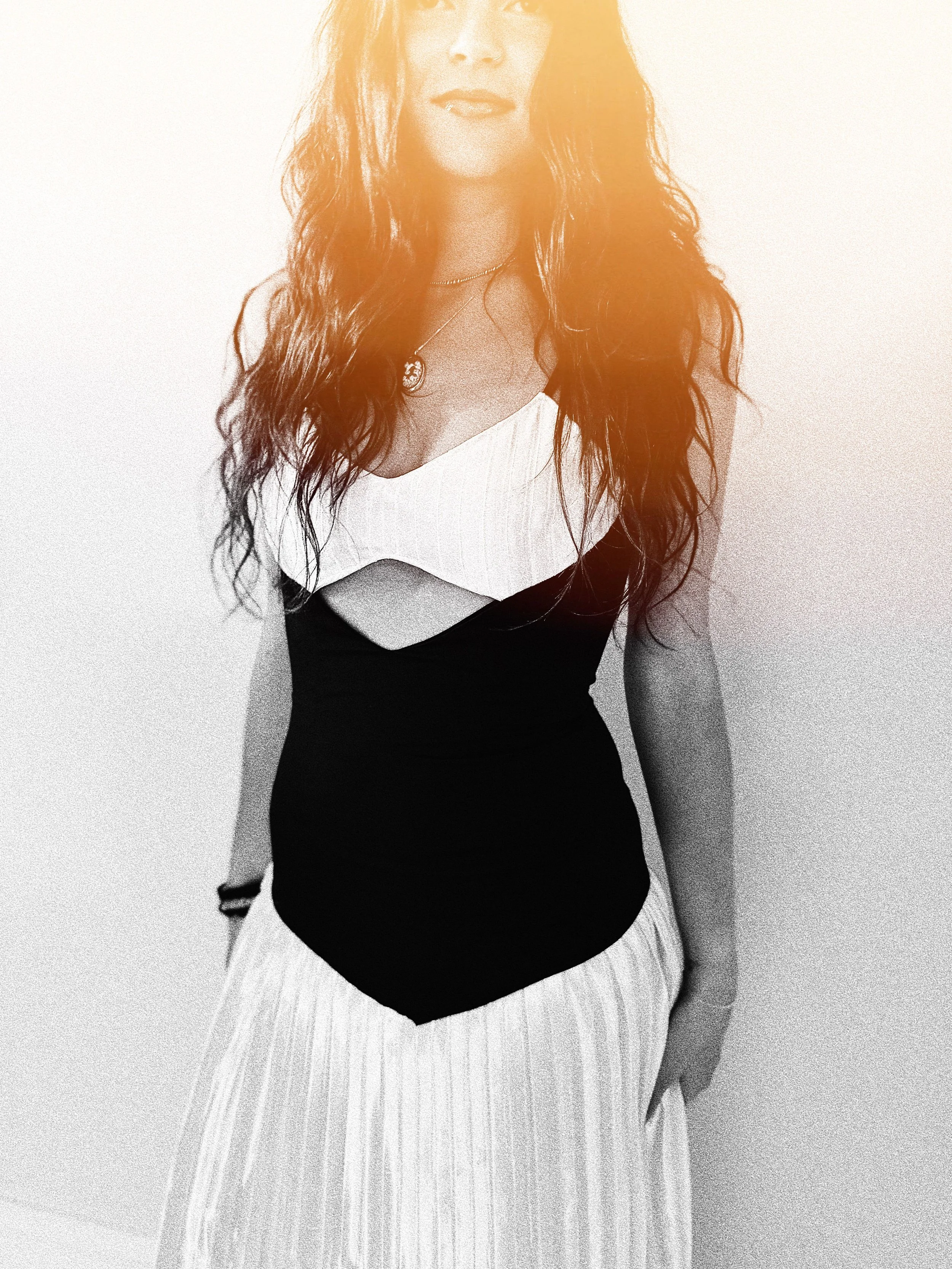 A black and white image of a woman with long wavy hair wearing a stylish top and a pleated skirt, standing against a plain background.