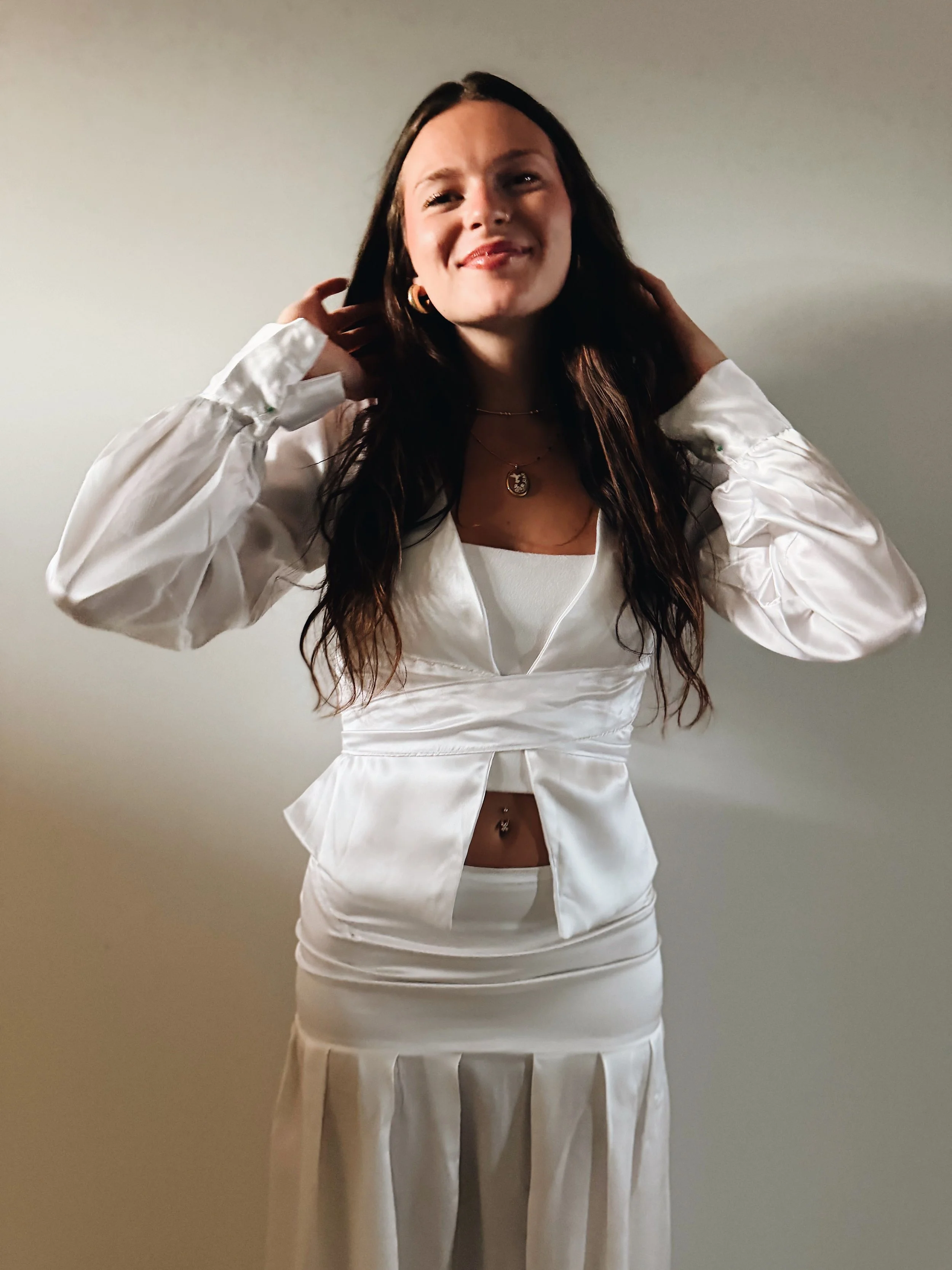 Woman with long dark hair wearing gold jewelry, a white satin blazer, matching skirt, and a white top, smiling and touching her hair, standing against a plain wall.