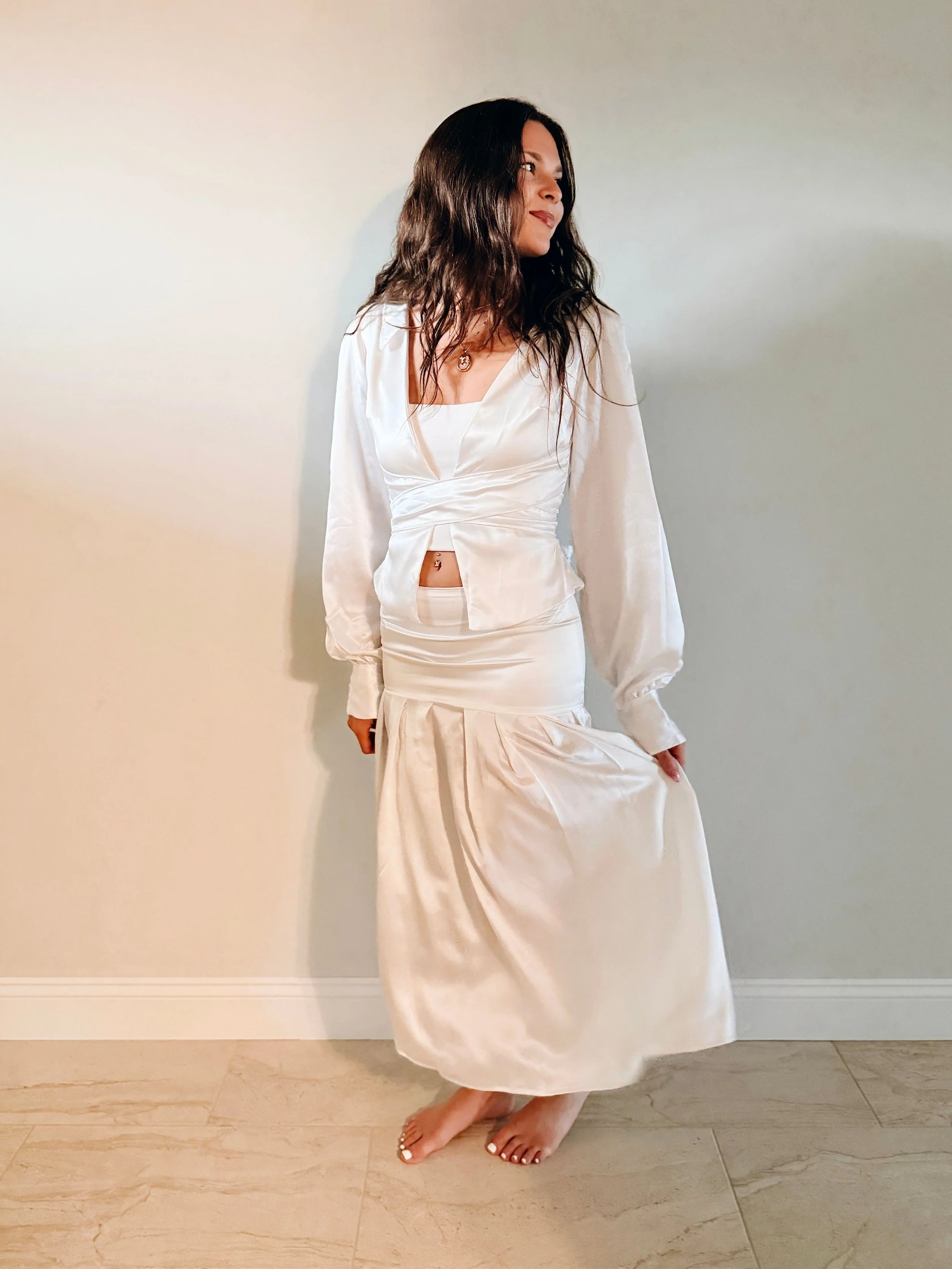 A woman with long, wavy dark hair wearing a white satin dress with a matching jacket and hat, standing barefoot on a light-colored tiled floor against a plain wall.