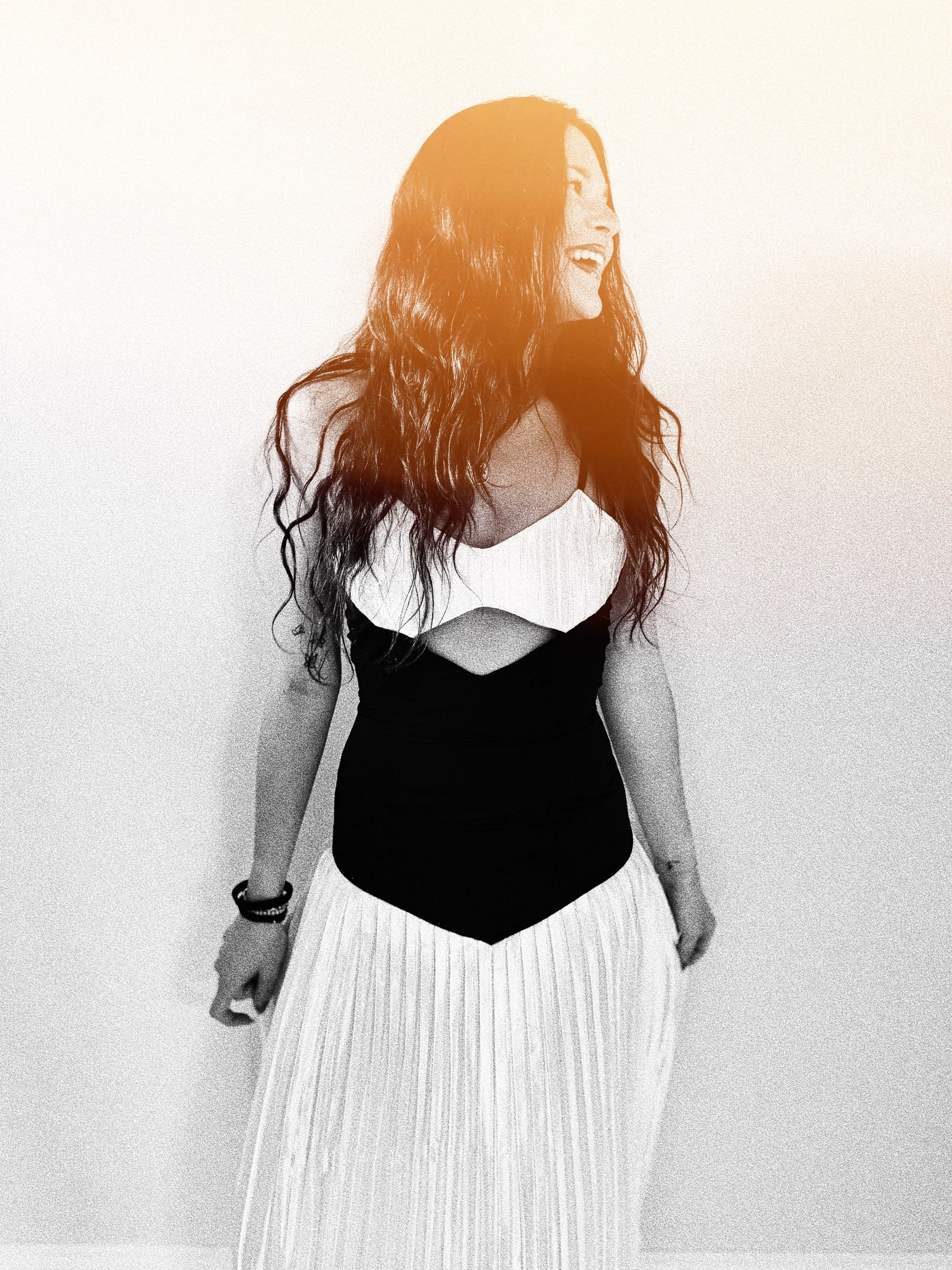 A woman with long, wavy hair laughing and looking to the side, wearing a sleeveless top and striped pants, standing against a plain background.