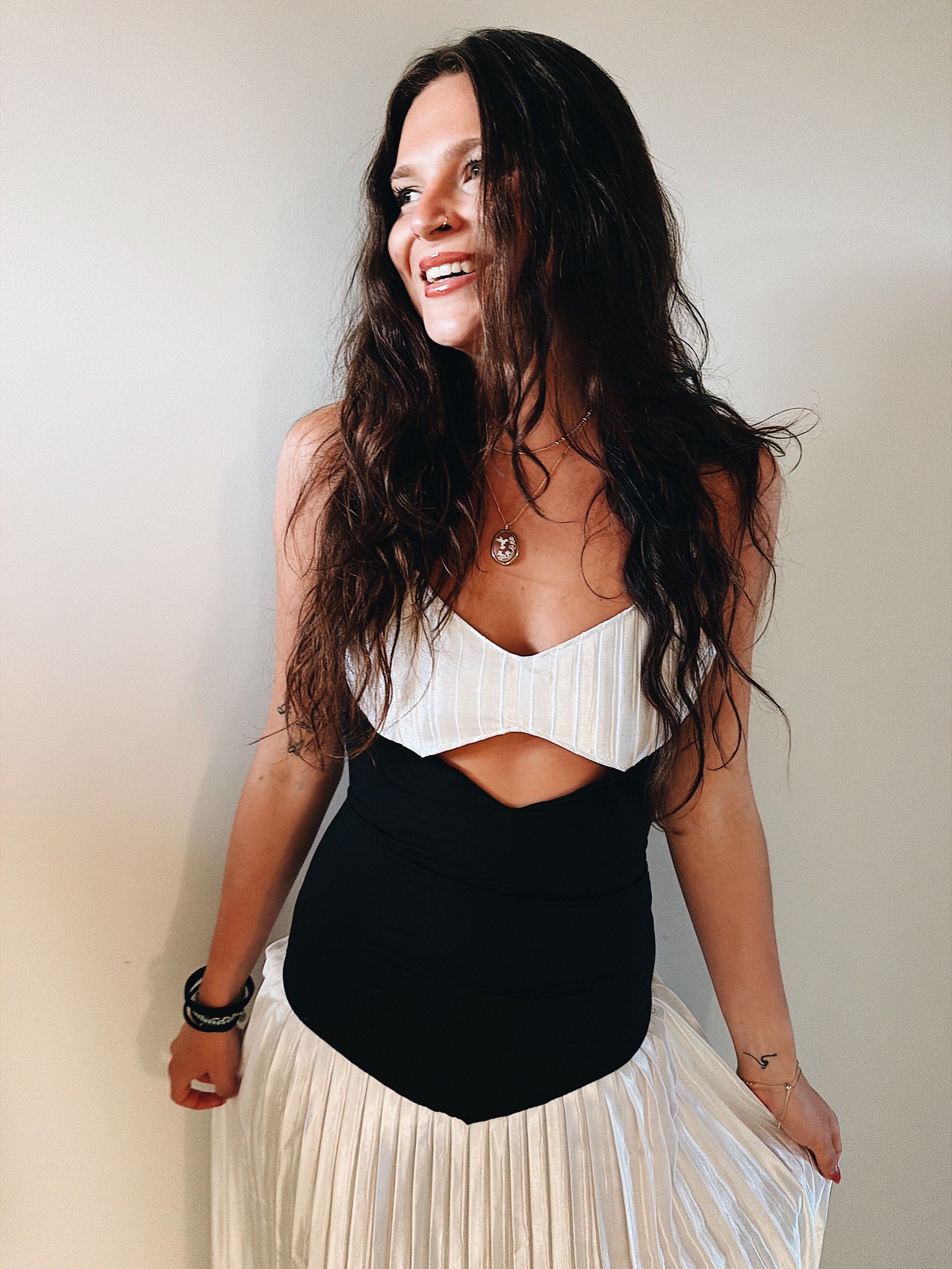 A woman with long, wavy dark hair smiling, wearing a layered necklace, a white pleated top, and a black dress with a cutout, standing against a plain light-colored wall.