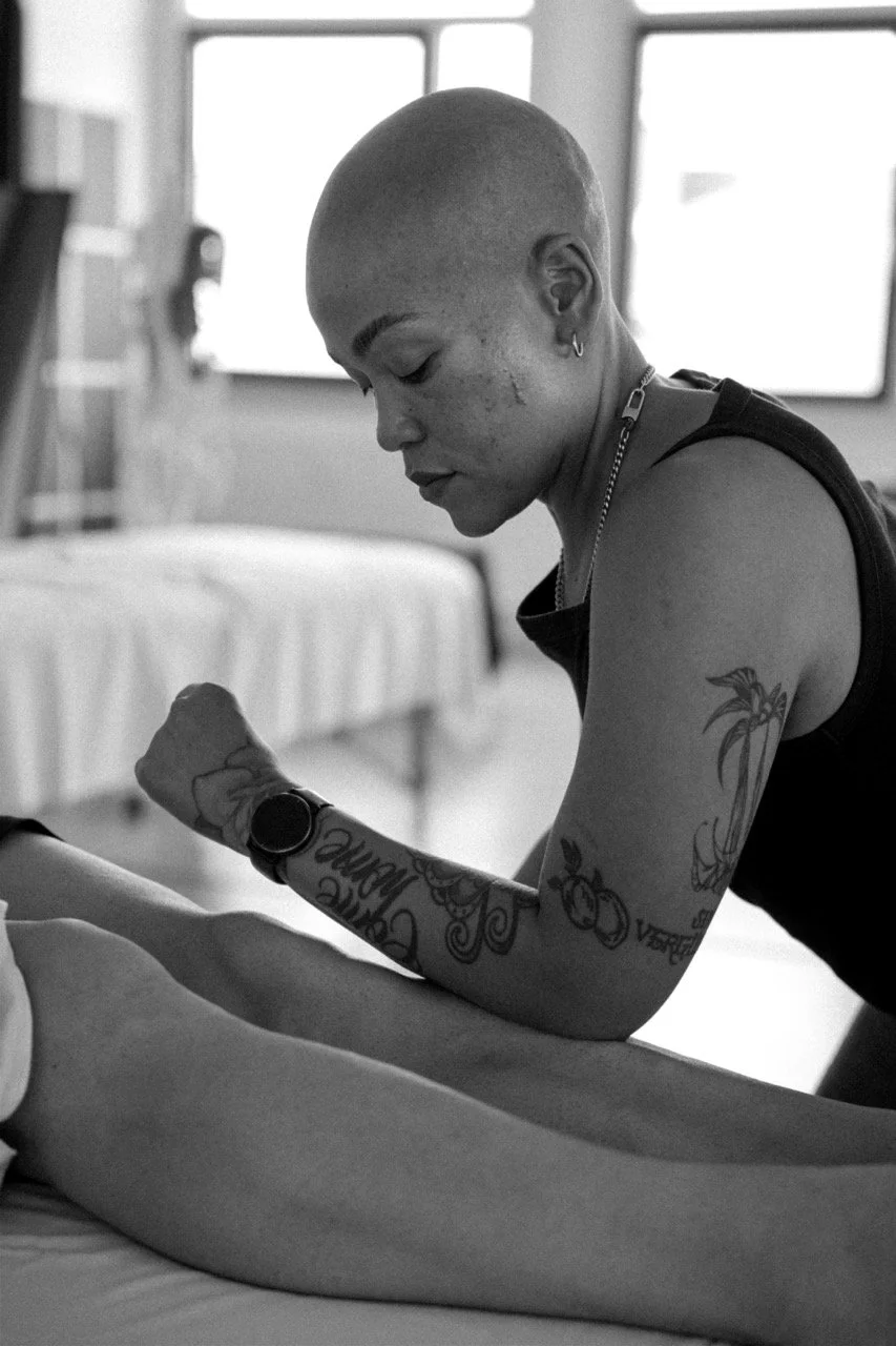 A woman with a shaved head and tattoos on her arm is showing her bicep and flexing in a room with large windows in the background.