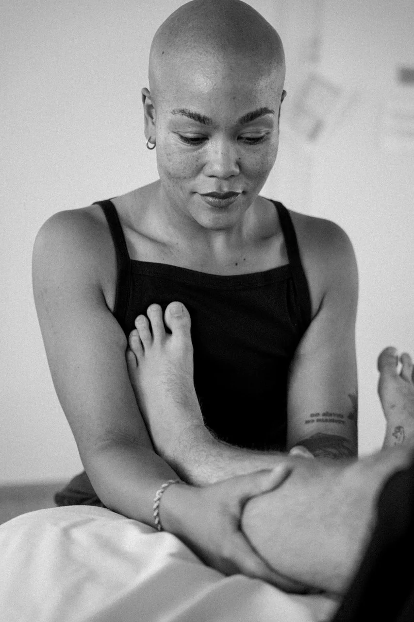 Black and white photo of a person with a shaved head and small earrings, wearing a sleeveless top, looking down at someone else's arm. The person appears to be comforting or supporting someone.