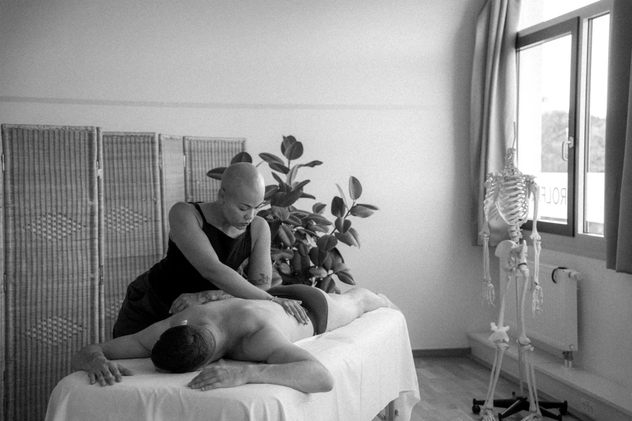 A person receiving a massage from a massage therapist in a treatment room with a skeleton model near the window and a folding privacy screen in the background.