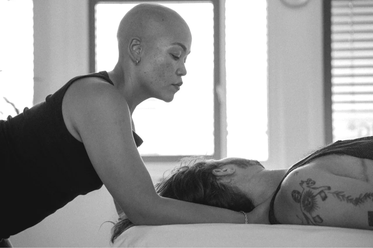 A woman is tending to a patient lying face down on a bed, likely giving a massage or medical treatment, in a well-lit room.