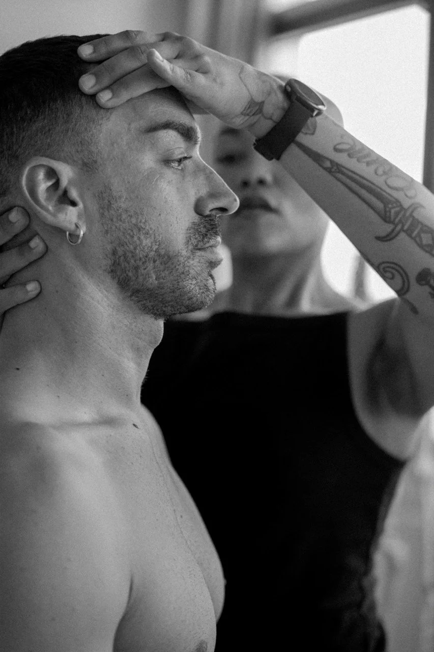 A person getting their makeup or face prepared with a hand placed on their forehead, while another person assists, with tattoos and watch visible on their arm, in a black and white photograph.