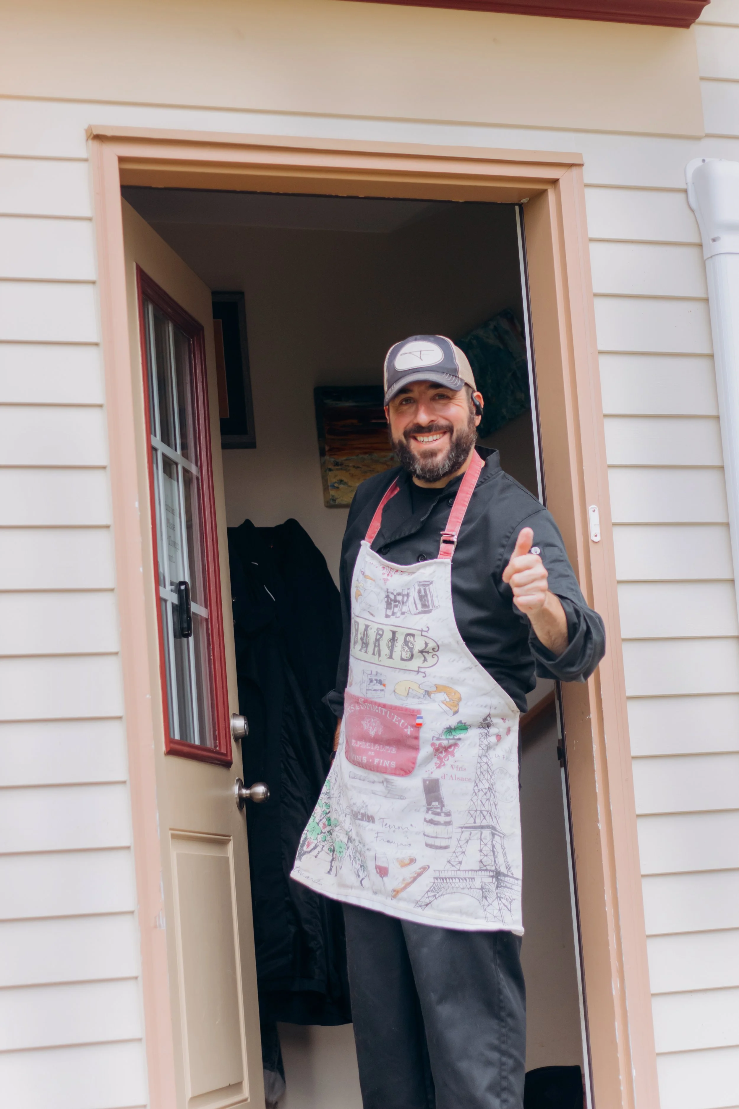 A man with a beard, smiling and giving a thumbs-up, standing in a doorway. He is wearing a baseball cap, a black shirt, and a colorful apron. The door leads into a cozy home with artwork visible inside.