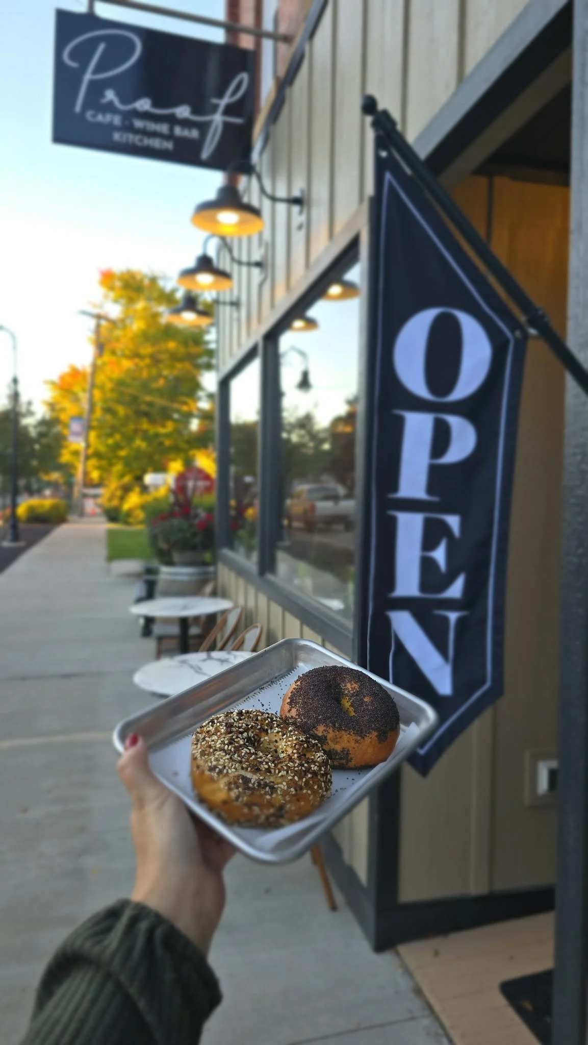 A person holding a tray of two donuts, one topped with sesame seeds and the other with poppy seeds, outside a bakery or cafe. The background shows a sidewalk, outdoor seating, and a sign that reads 'Open'