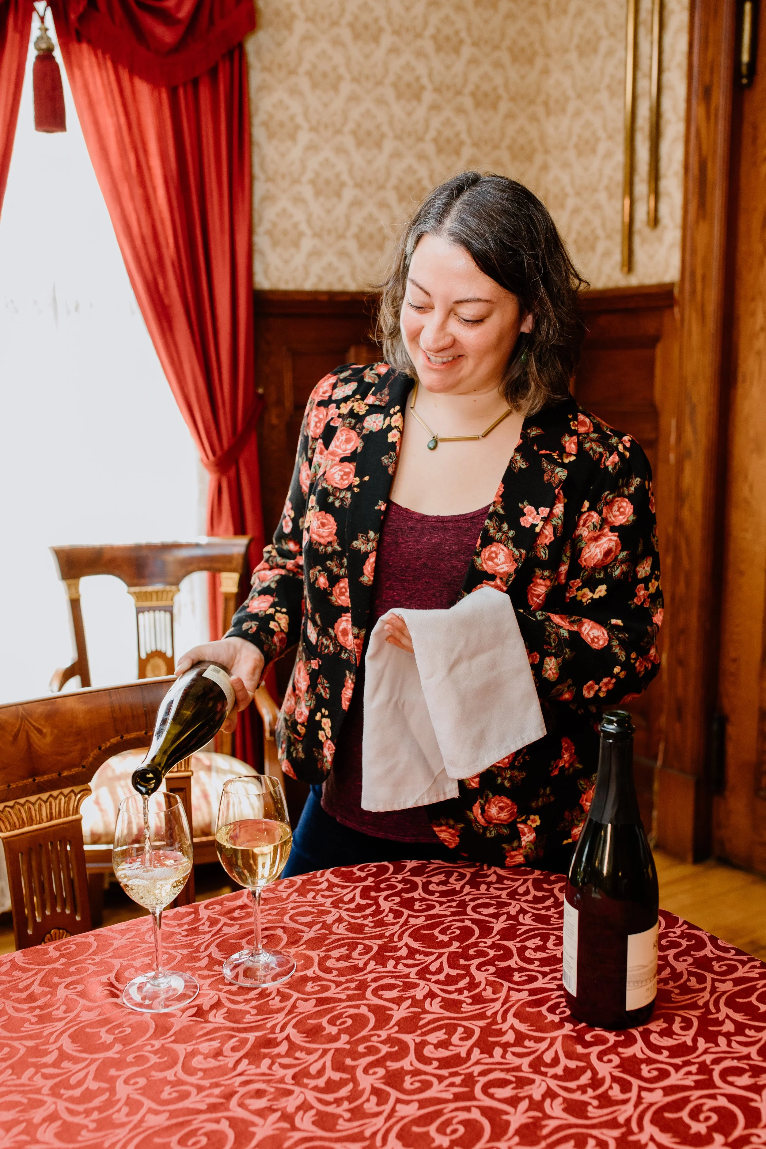 A woman in a floral blazer pouring white wine into a glass at a table with a red patterned tablecloth, with wine bottles and glasses on the table in a warmly decorated dining room.