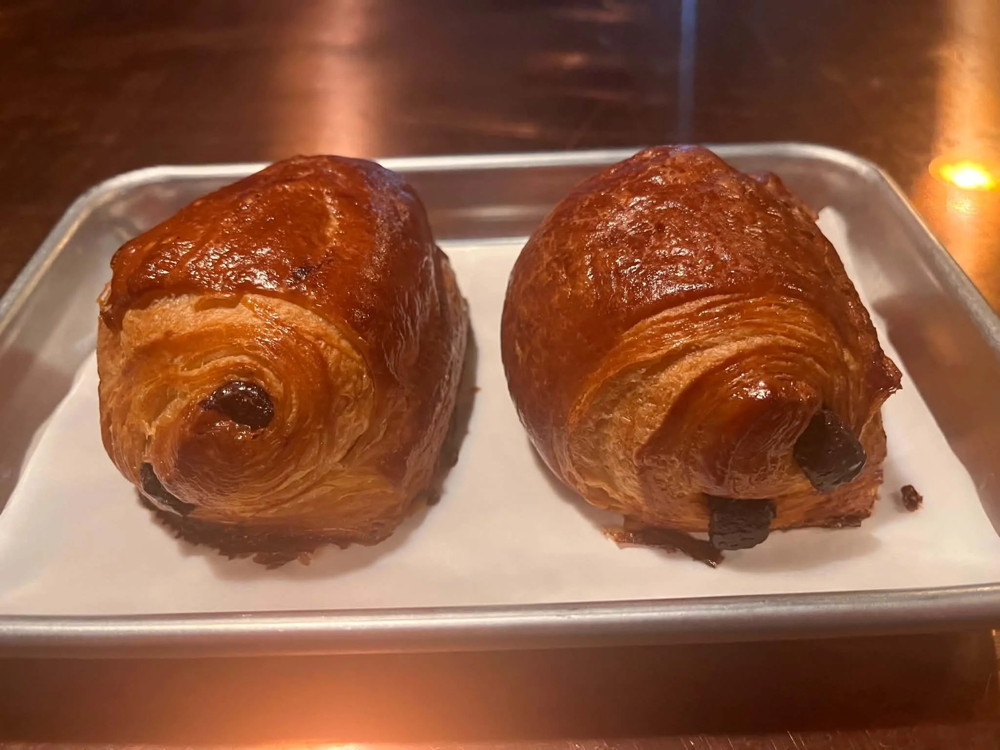 Two golden-brown croissants with a glossy finish, placed on a metal tray.