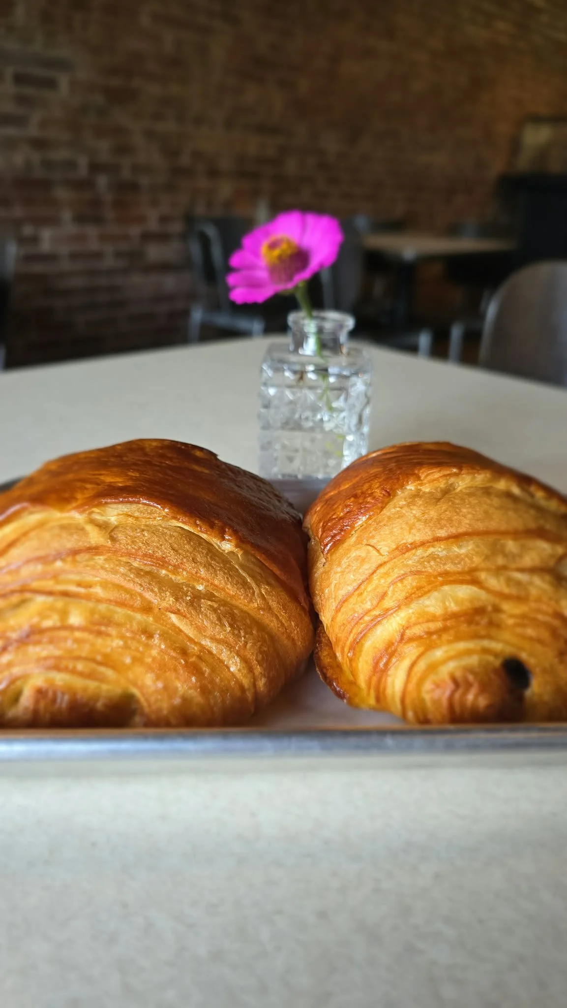 Two golden-brown croissants on a white tray in the foreground with a small vase holding a pink flower behind them on a table inside a restaurant with brick walls.