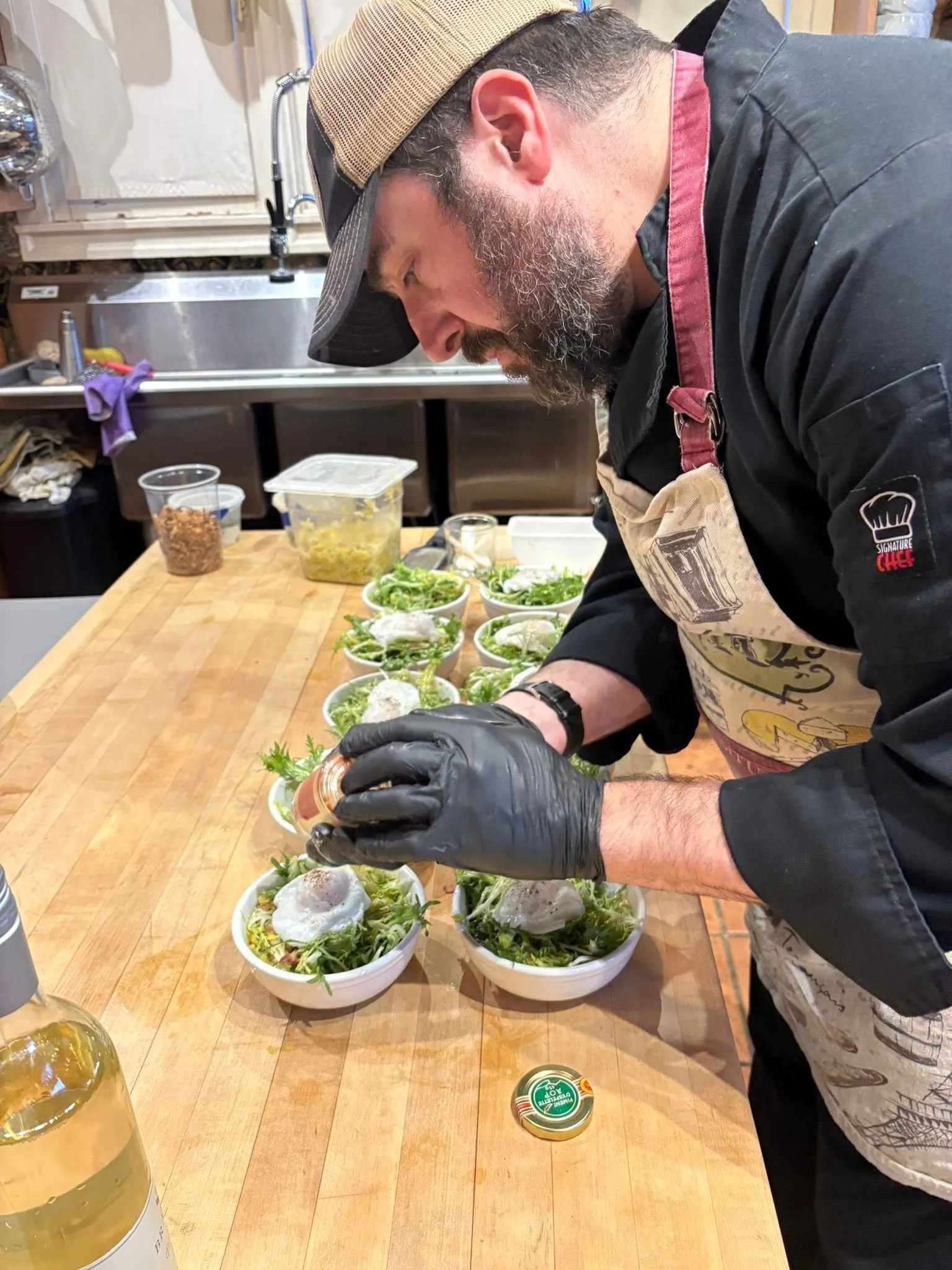 A man wearing a beige cap, black gloves, a black apron, and a dark shirt preparing salads in bowls on a wooden kitchen counter. The bowls contain green lettuce, topped with poached eggs, and some toppings. In the background, there are various kitchen items, a container of chopped yellow vegetables, and a purple cleaning cloth.