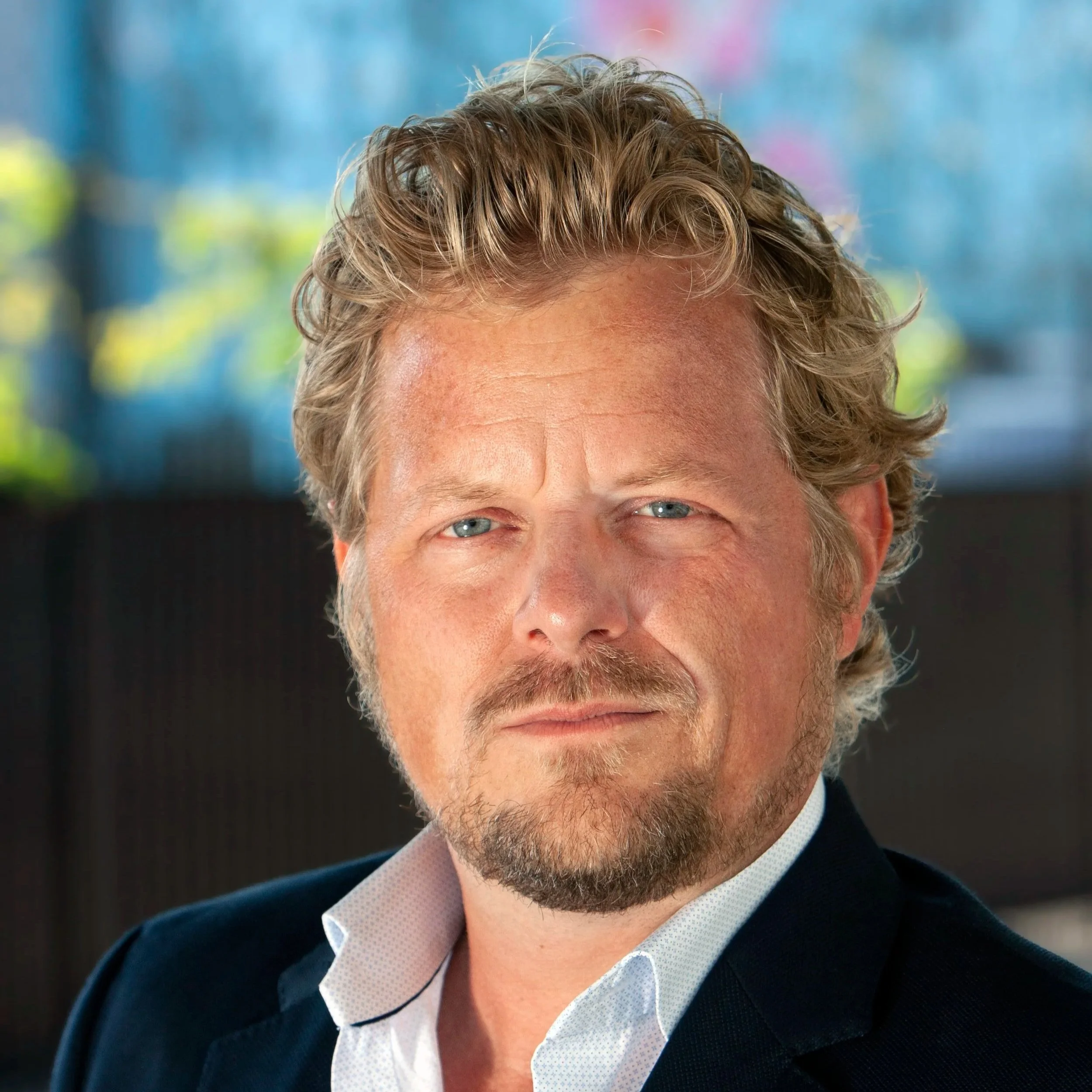 Close-up of a man with curly blond hair, blue eyes, and a short beard, wearing a white shirt and a dark suit jacket