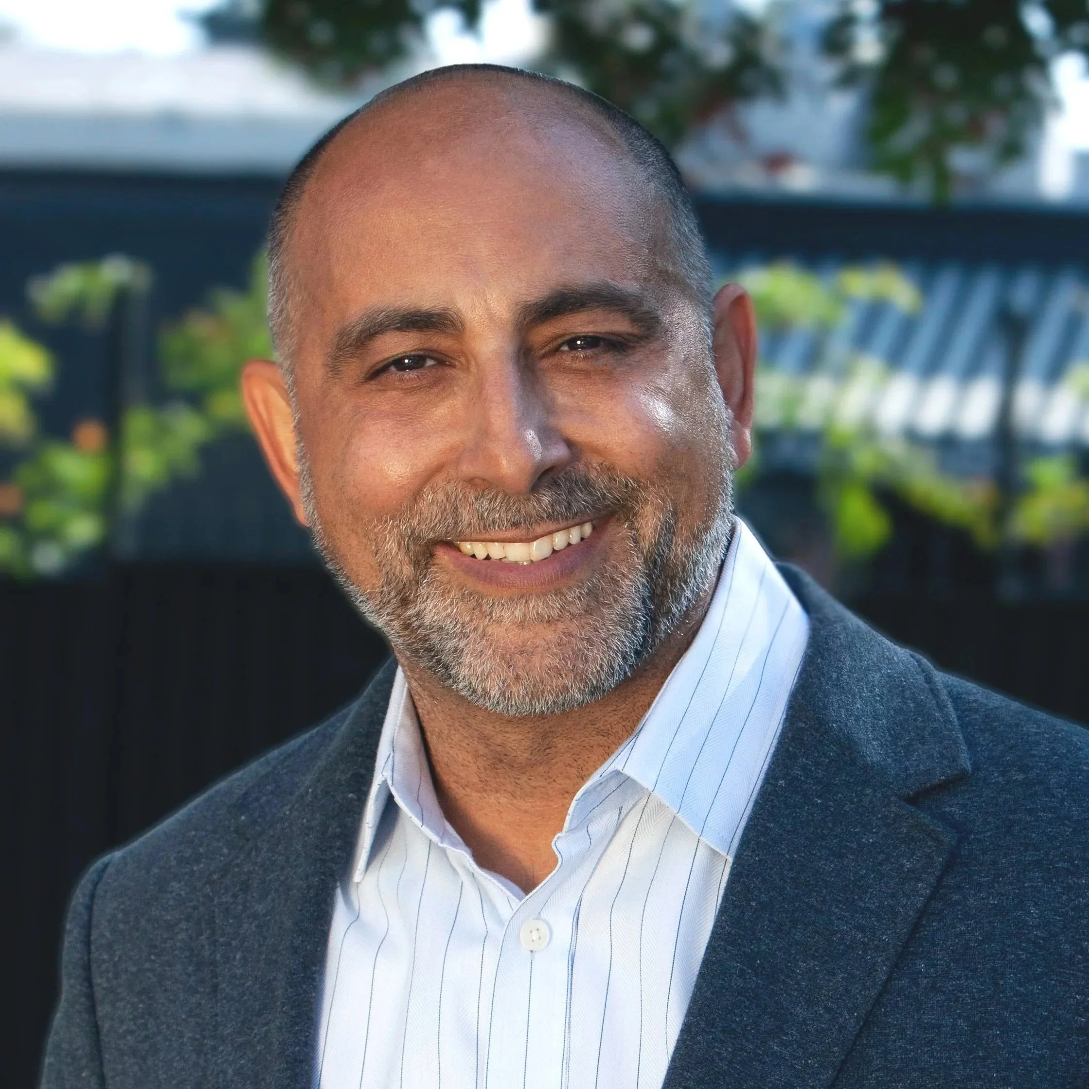 Close-up of a smiling man with a beard, wearing a blue suit and a white shirt with thin blue stripes, outdoors with blurred greenery in the background.
